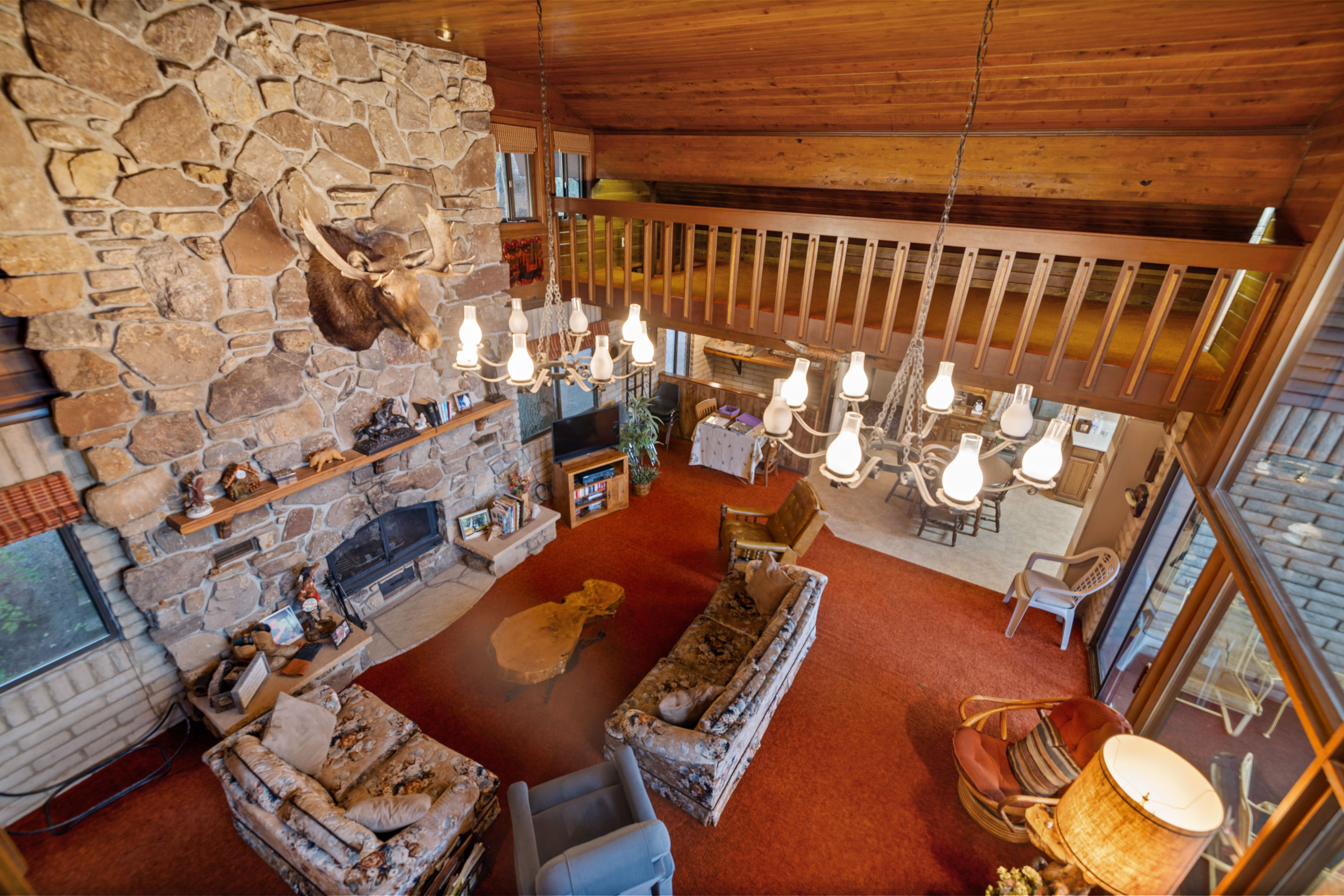 Great room featuring a floor-to-ceiling stone fireplace, wooden vaulted ceiling, a loft with wood railing, and an exposed brick accent wall
