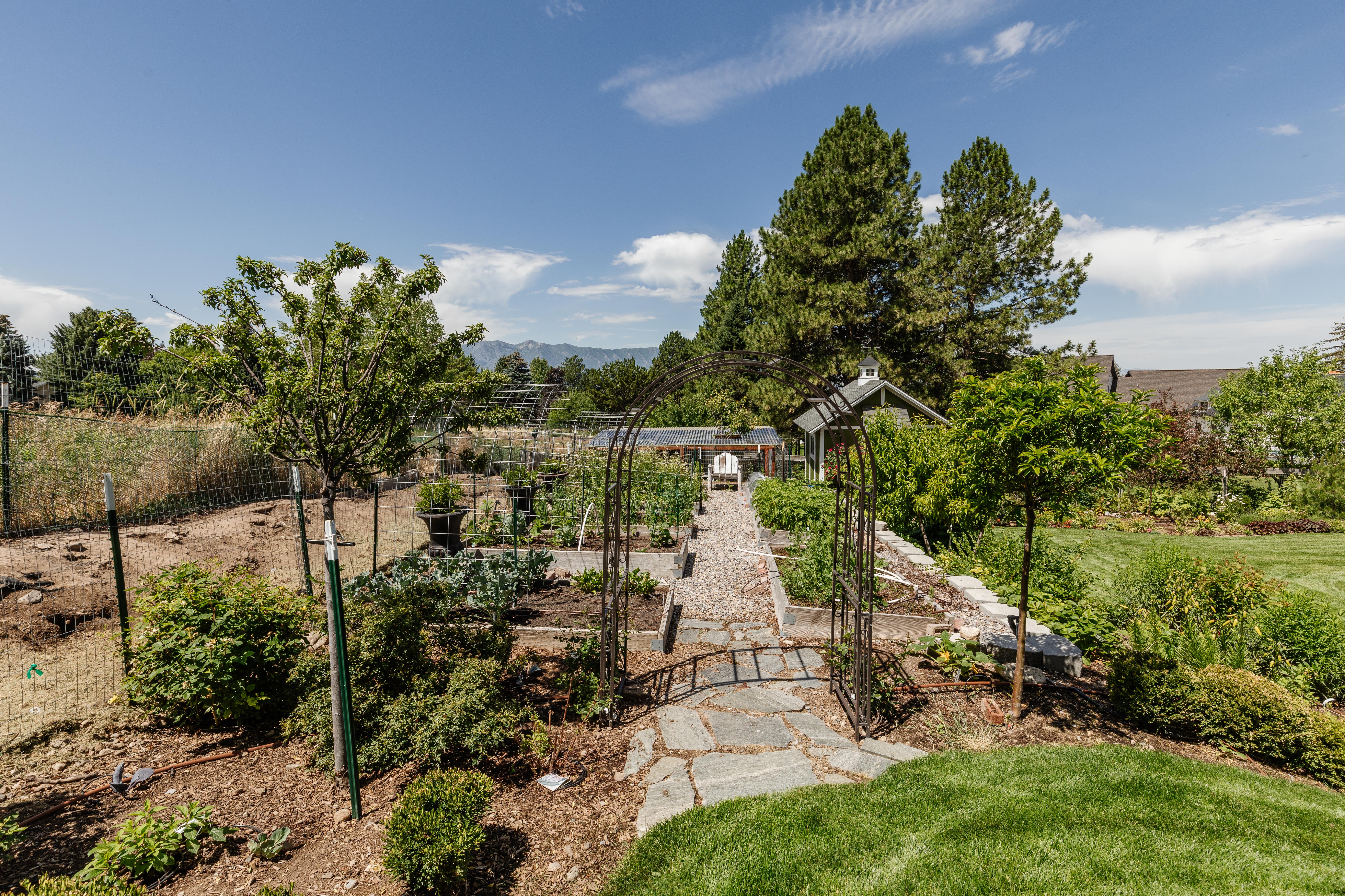 Expansive garden area featuring a stone pathway, raised garden beds, a decorative metal archway, and mature trees