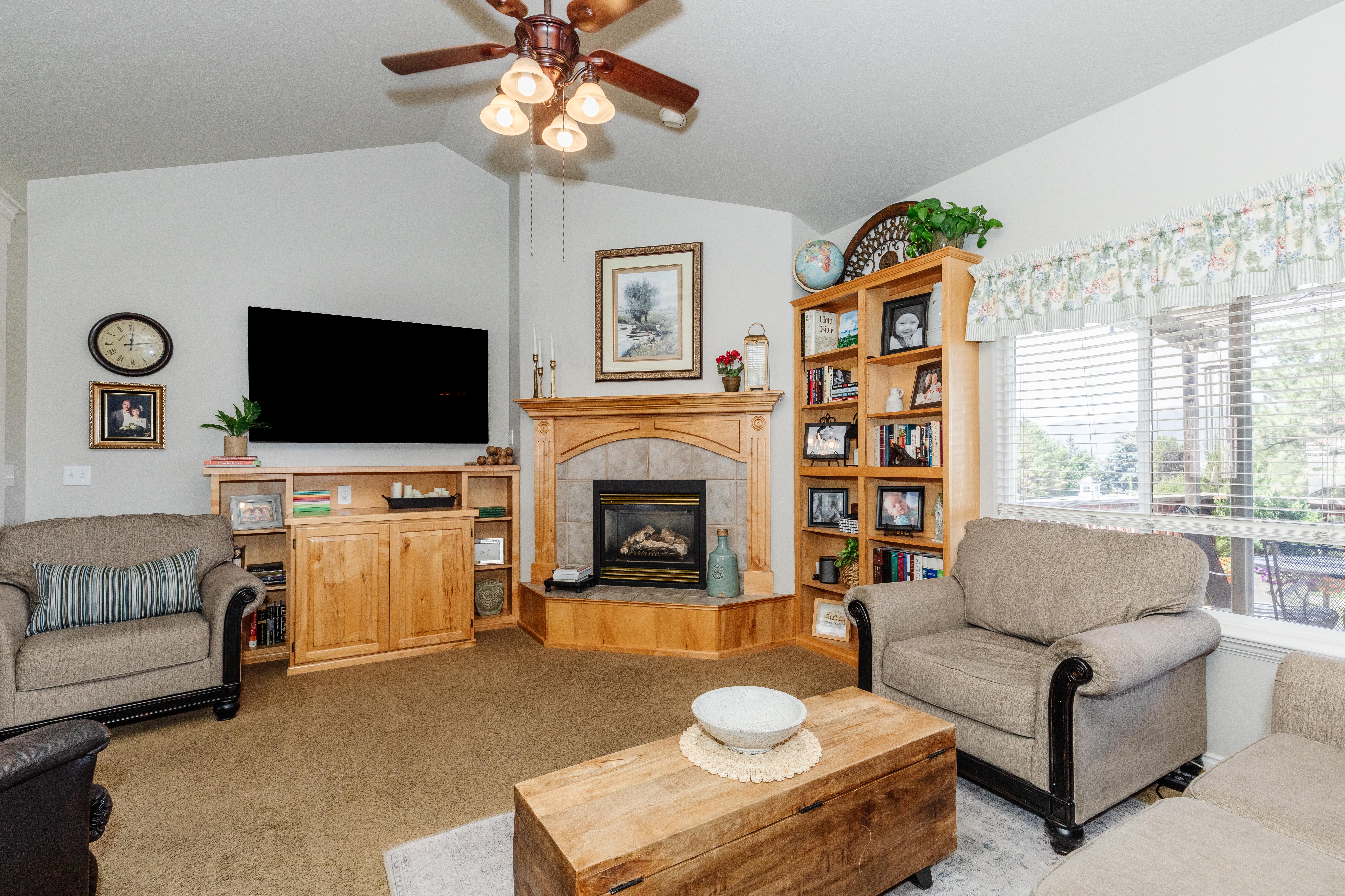 Vaulted ceiling with a ceiling fan light fixture, corner gas fireplace with a wood mantel and stone surround, built-in shelving, and a large window with blinds