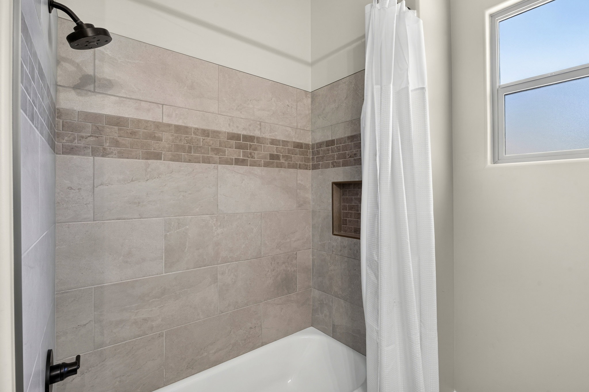 Bathtub and shower combination featuring a dark bronze shower head and faucet, light-toned wall tiling with a mosaic accent band, and a built-in shower niche