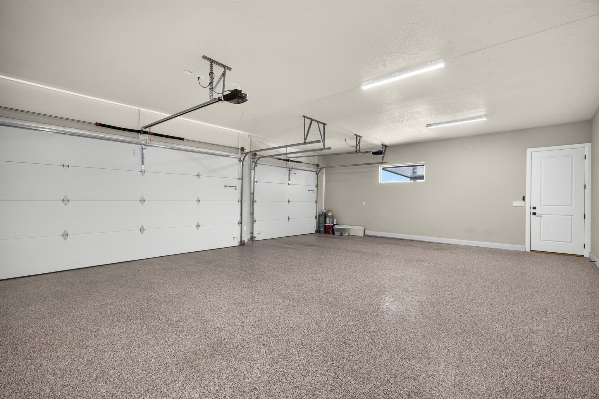 Spacious garage featuring a durable speckled epoxy floor, two overhead garage doors, overhead lighting, a white panel door, and a horizontal window