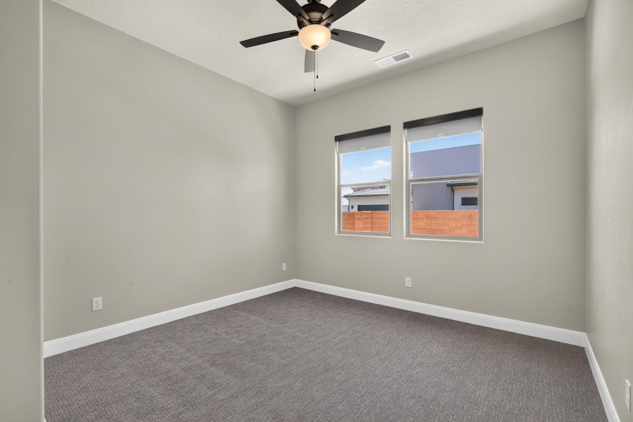 Neutral-toned room featuring a black ceiling fan with integrated lighting, two windows with roller shades, and plush grey carpeting