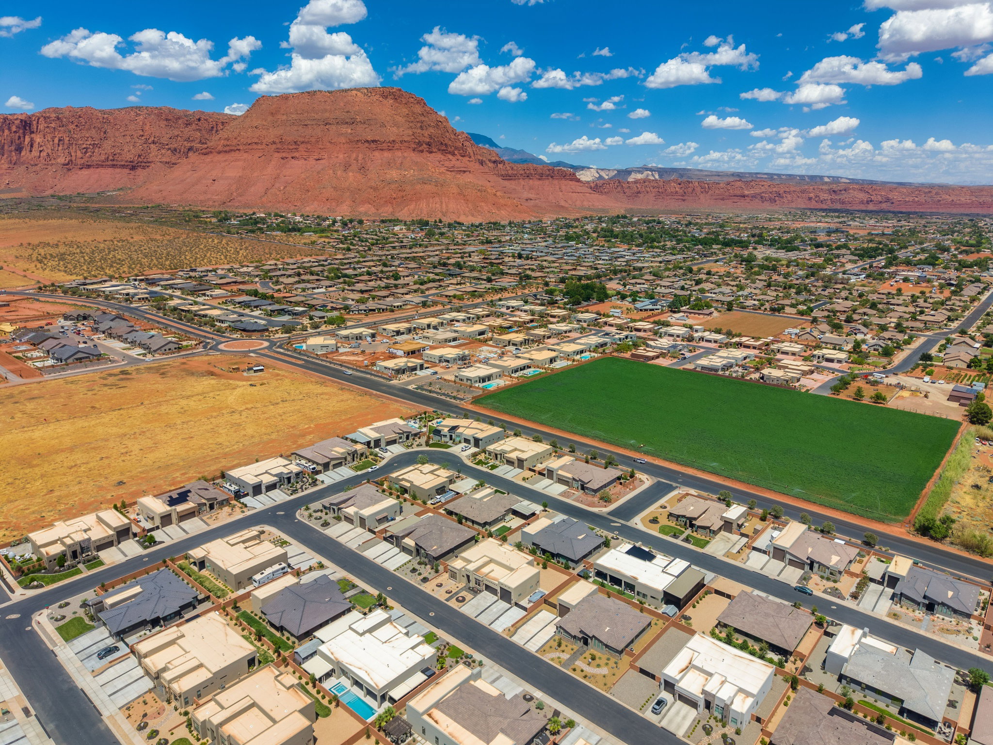 Expansive aerial perspective showcasing a residential development with varied architectural styles, a vibrant green field, and natural red rock formations in the background
