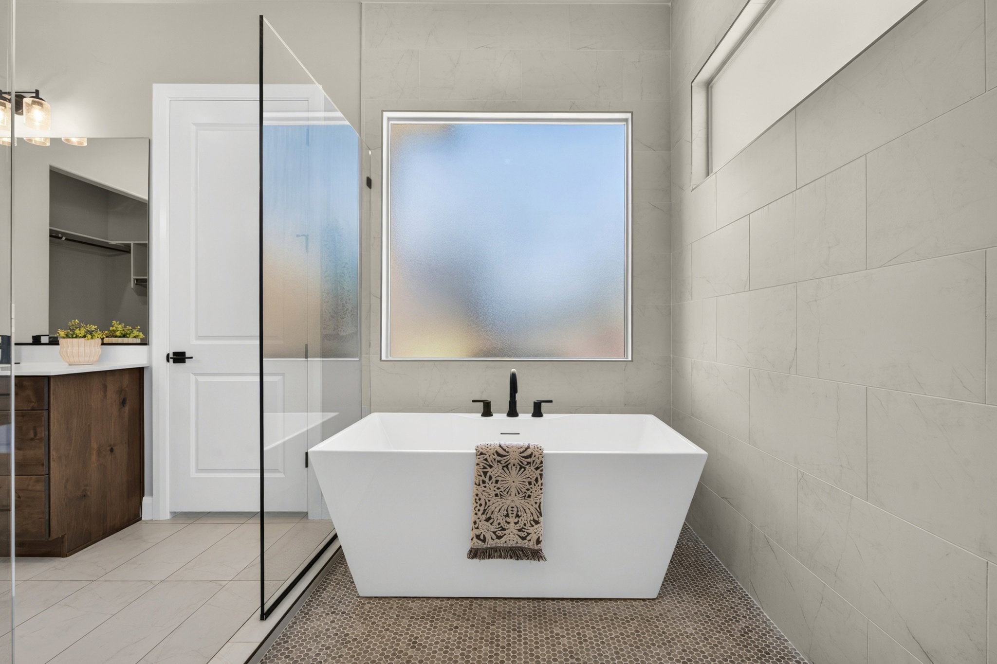 Freestanding soaking tub with matte black fixtures, set against large format wall tiling and a frosted window