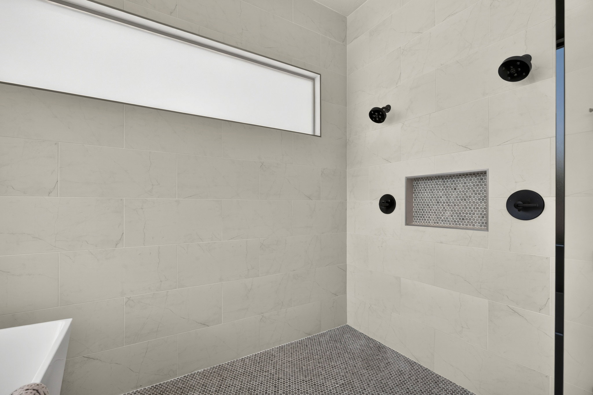 Spacious shower with large format wall tiling, matte black fixtures, an integrated tiled niche, and a mosaic tile floor