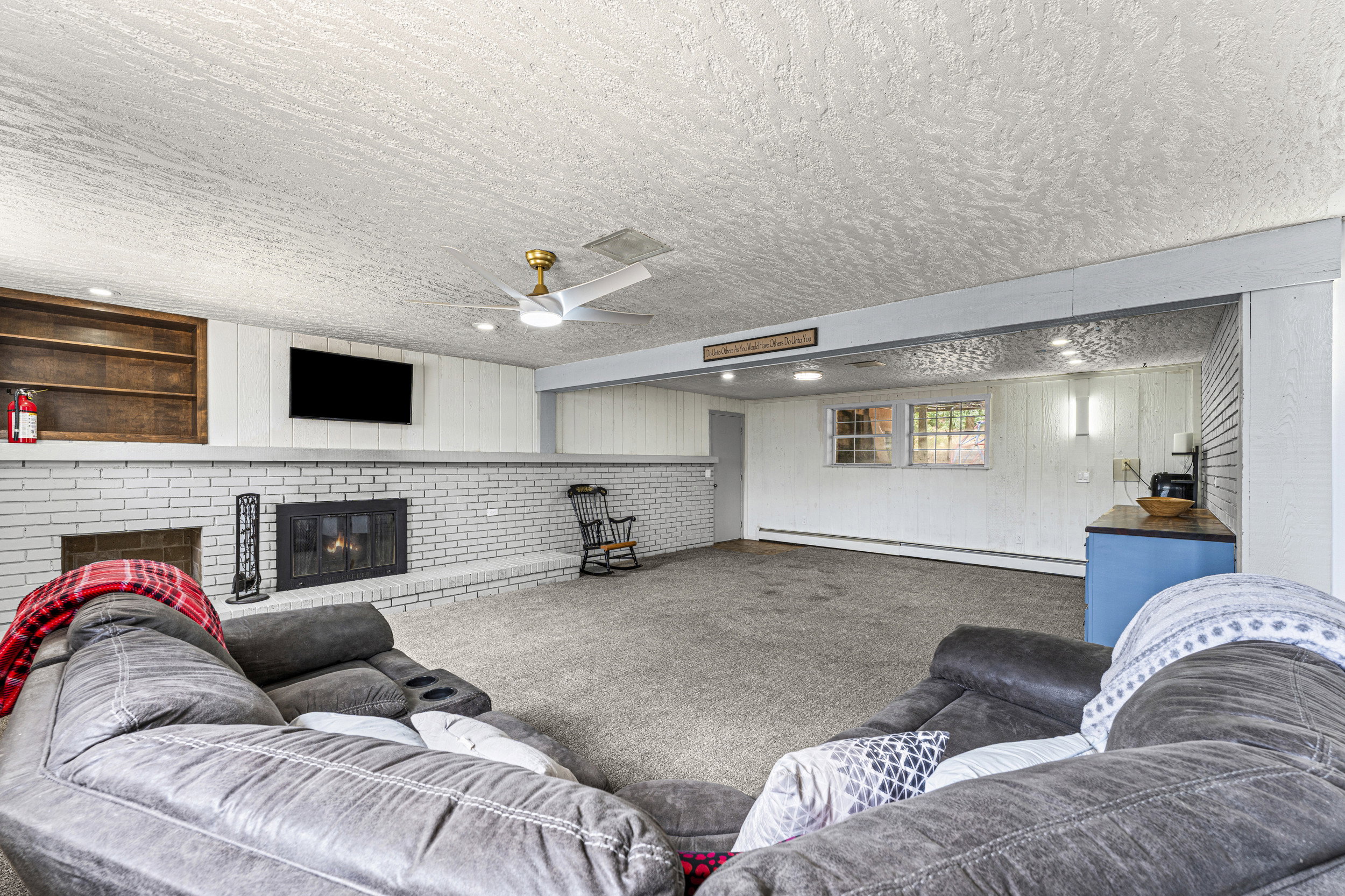 Spacious lower-level room featuring a white brick fireplace, recessed lighting, ceiling fan, and built-in shelving