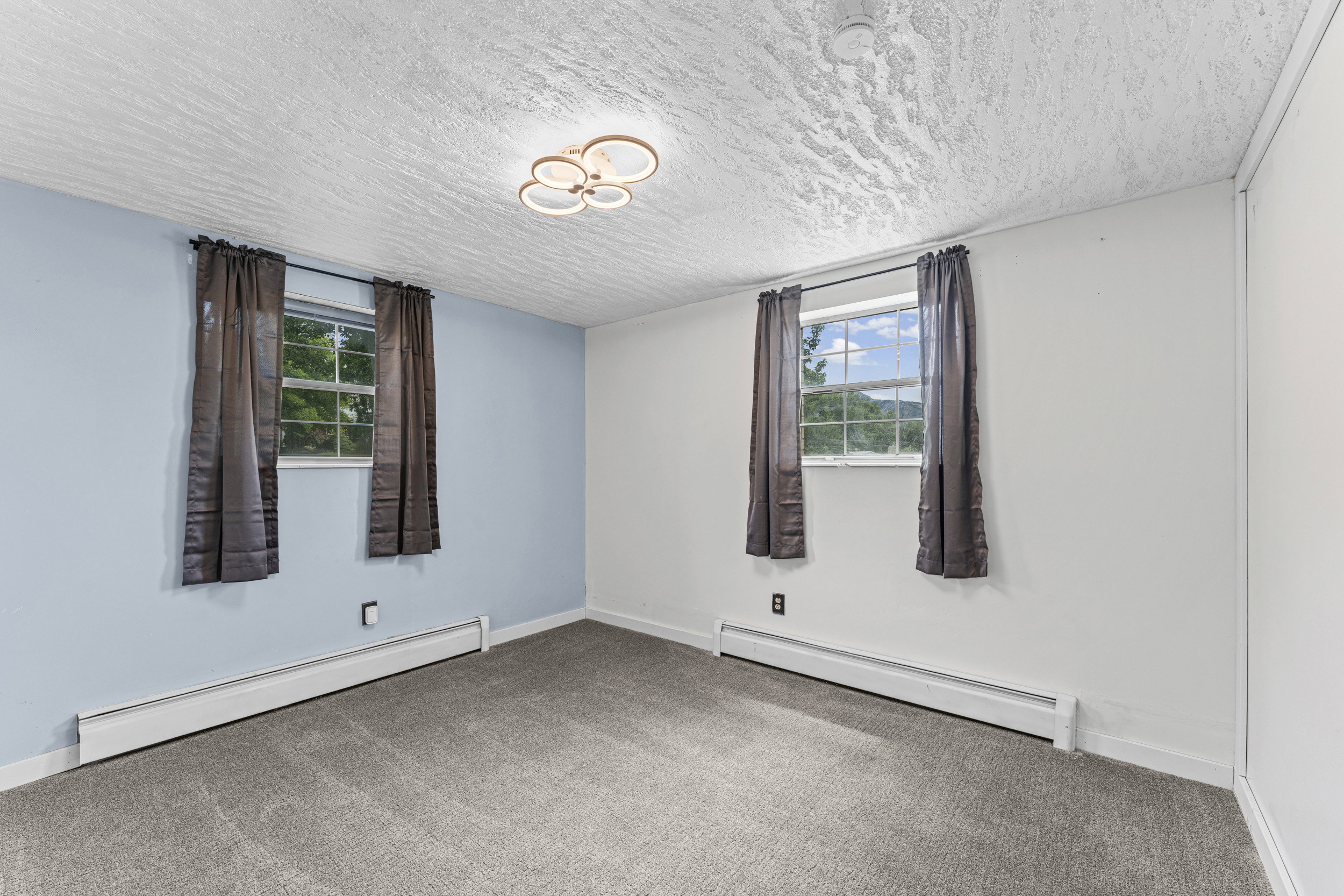 Carpeted room featuring two windows with curtain rods, baseboard heating, a modern ceiling light fixture, and a textured white ceiling