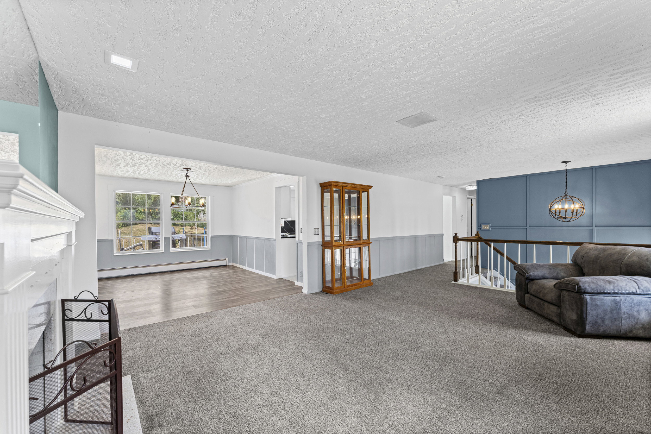 Spacious open-concept living area featuring a white fireplace with an ornate screen, gray carpeting, a decorative blue accent wall with paneling, and a contemporary spherical pendant light fixture