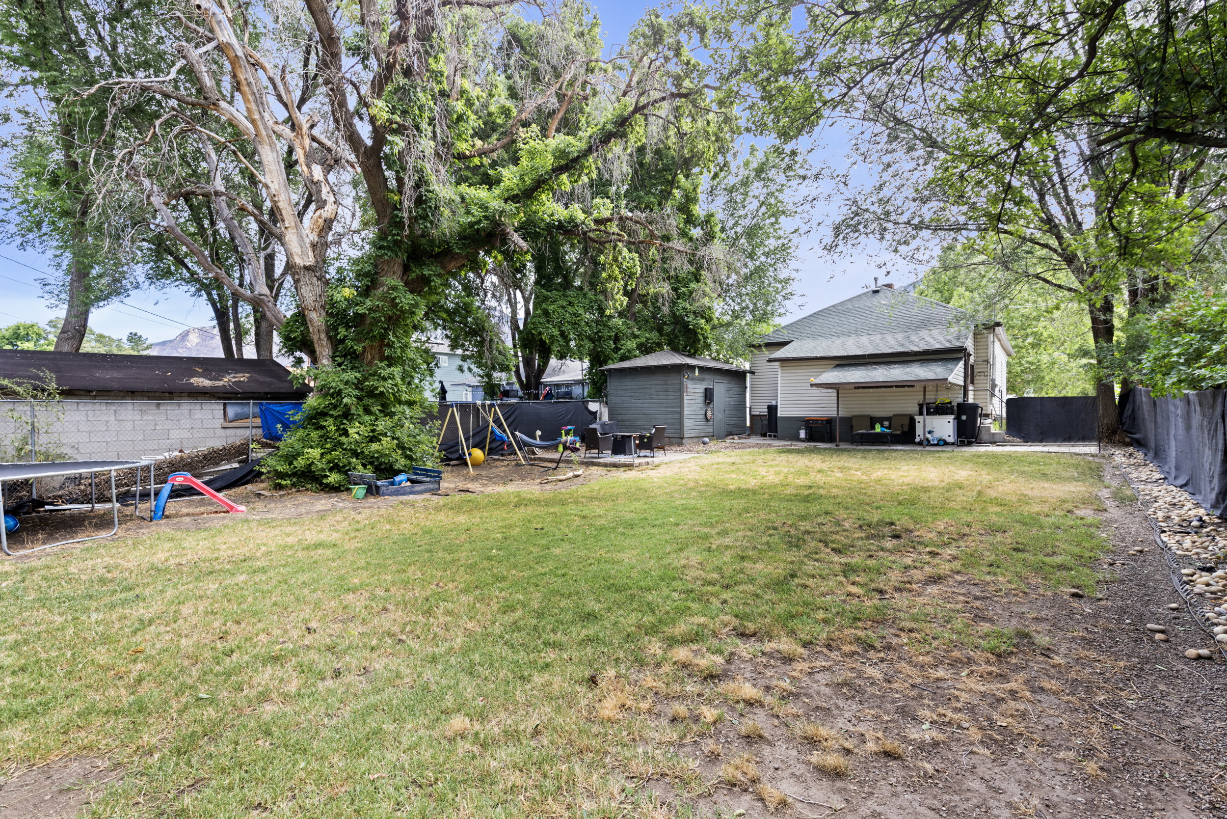 Expansive grassy yard area featuring mature trees, a permanent storage shed, and a covered patio area on the rear of the property