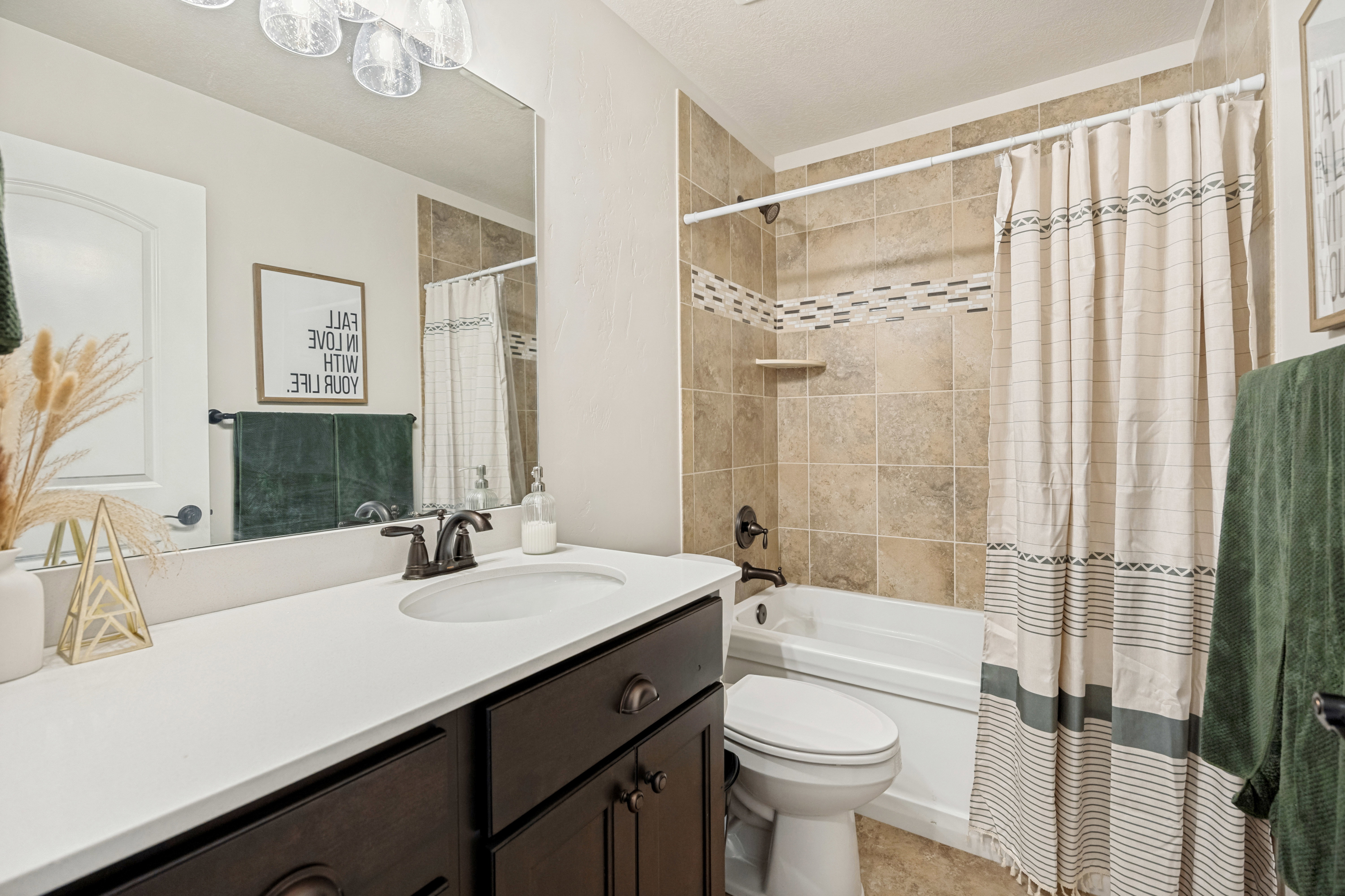 Bathroom featuring a vanity with dark wood-finish cabinetry and a white countertop, an undermount sink with a dark bronze faucet, and a shower-over-tub combination with tiled walls and an accent tile band