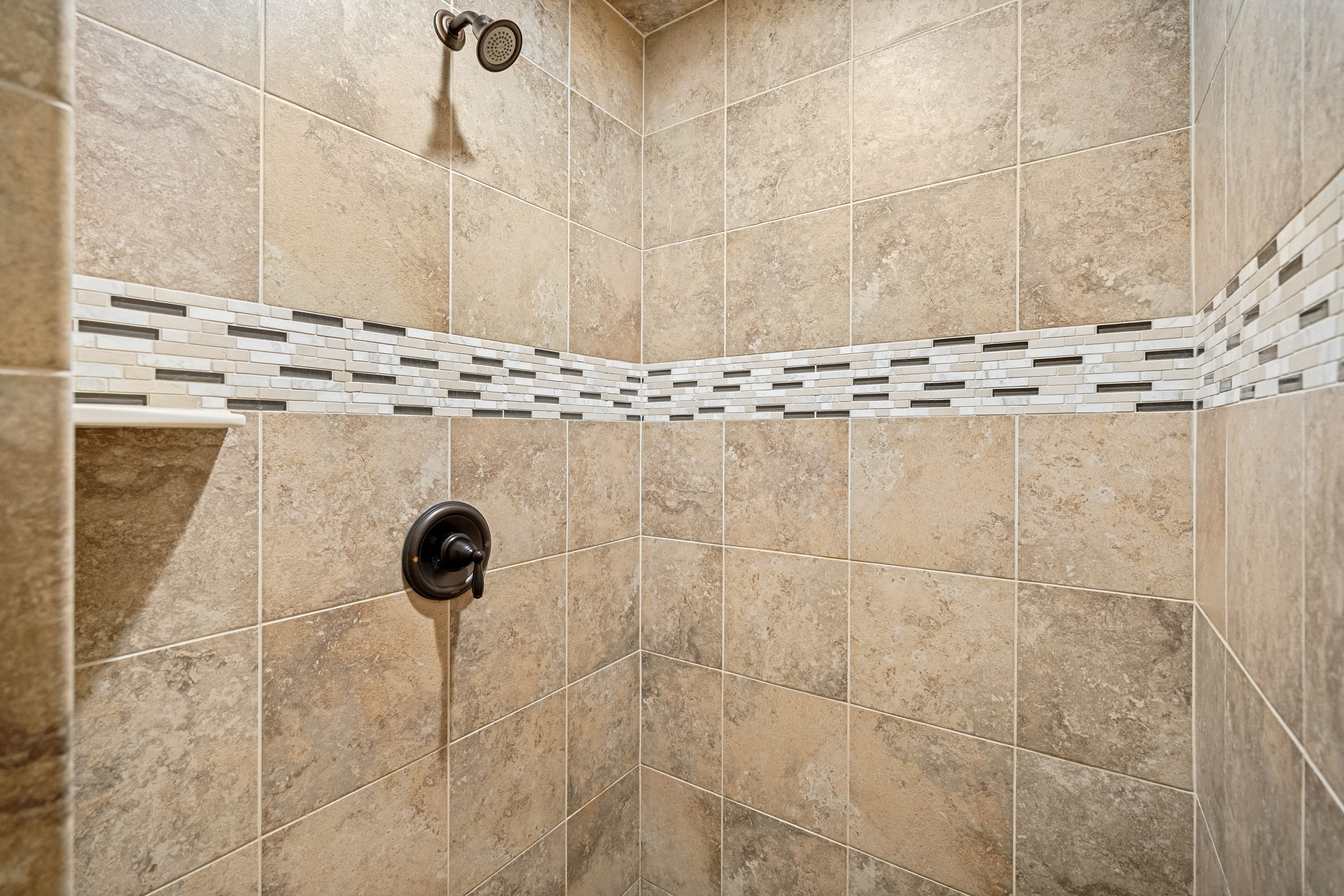 Shower enclosure featuring large format stone-look tile, decorative mosaic tile accent band, oil-rubbed bronze shower head, and oil-rubbed bronze shower control
