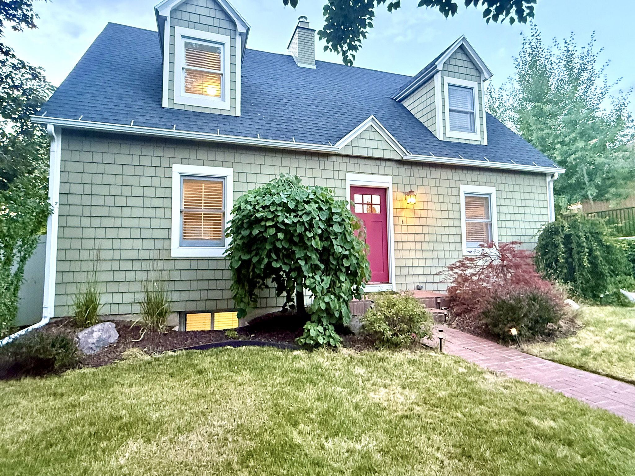 Dormered dwelling with green shingle siding, a red front door, and a brick pathway