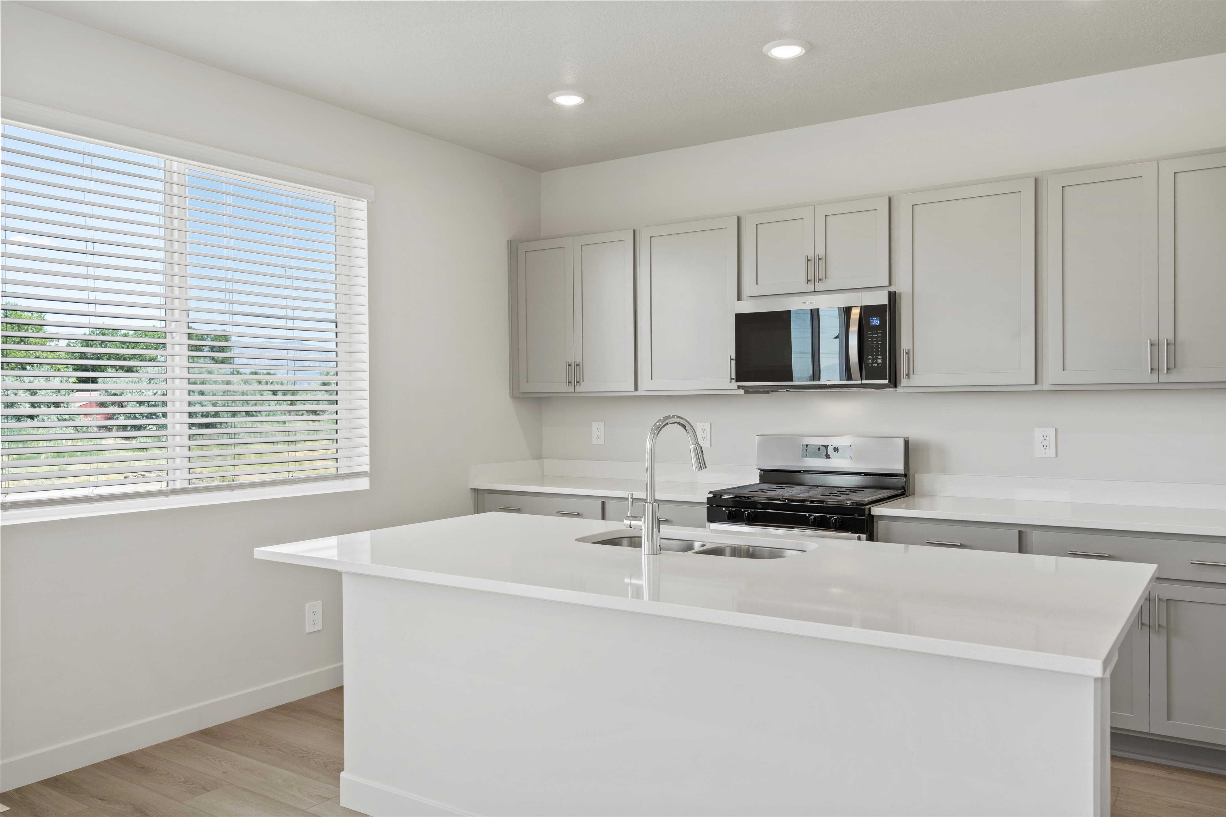 **DISCLAIMER** Interior package may vary. Open-concept kitchen featuring an island with an undermount sink and gooseneck faucet, light grey cabinetry, white countertops, and wood-finish flooring