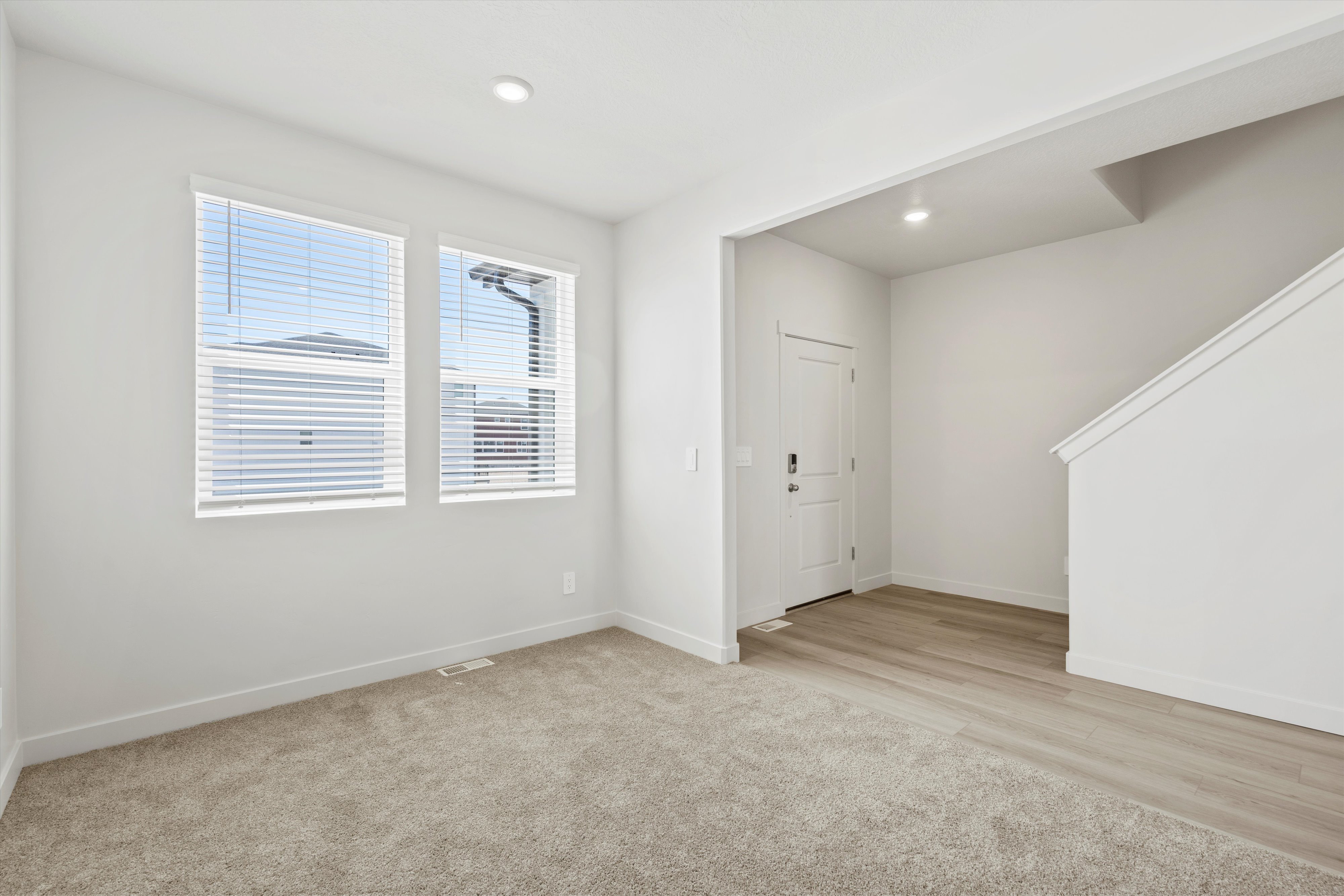 **DISCLAIMER** Interior package may vary. Living area featuring two large windows with blinds, recessed lighting, neutral paint, and a combination of light-colored carpeting and wood-finish flooring