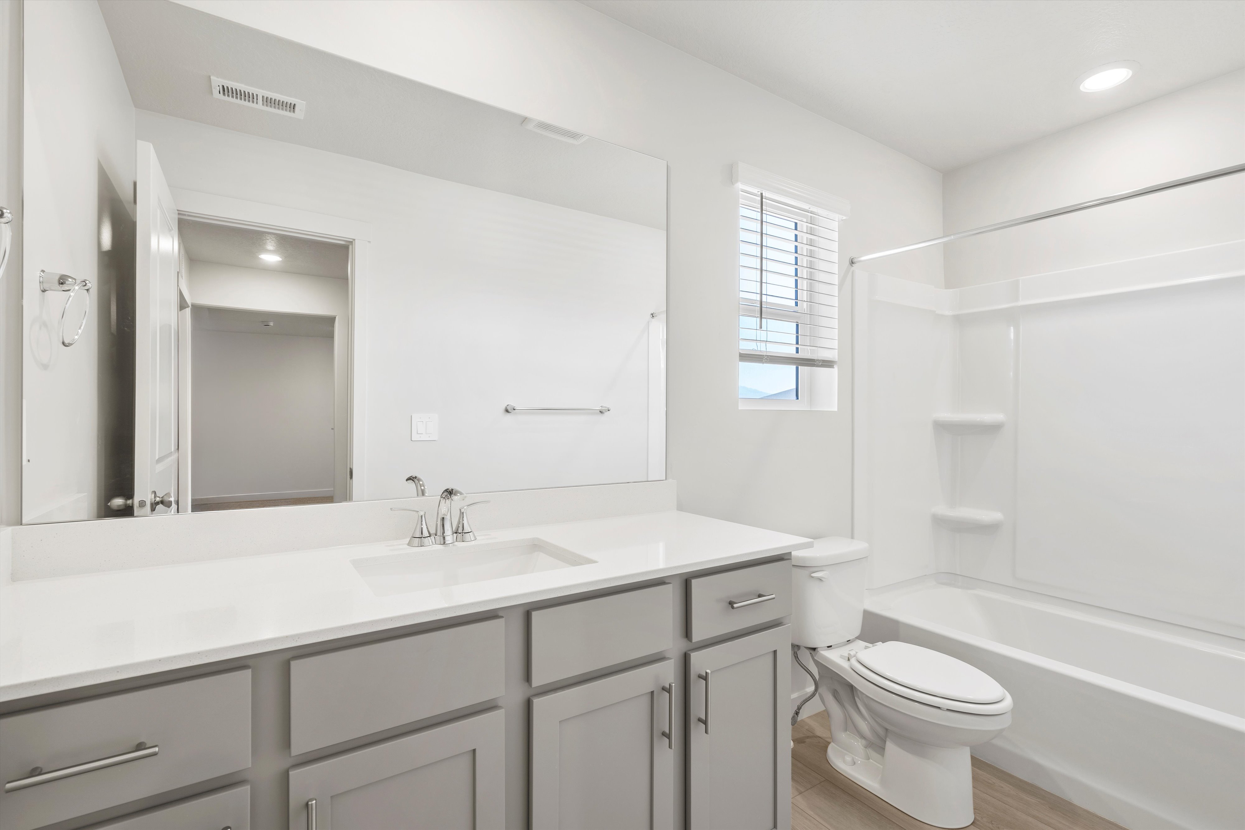 **DISCLAIMER** Interior package may vary. Bathroom featuring a single vanity with light gray cabinetry and a white countertop, chrome fixtures, a large mirror, a shower-over-tub combination with white surround, and wood-finish flooring