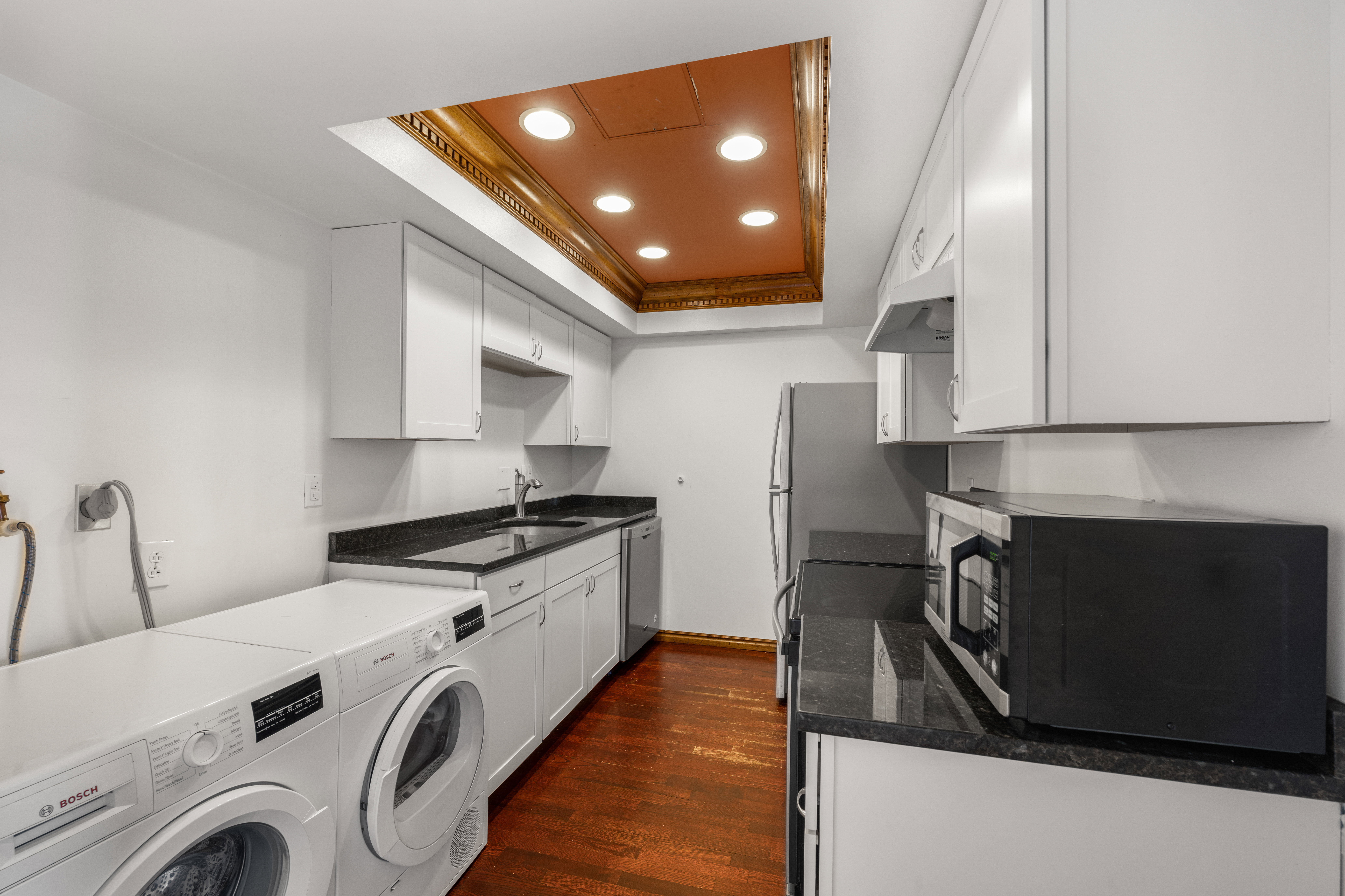 Integrated laundry and kitchen area featuring white cabinetry, dark granite-style countertops, wood-finish flooring, and recessed ceiling lighting