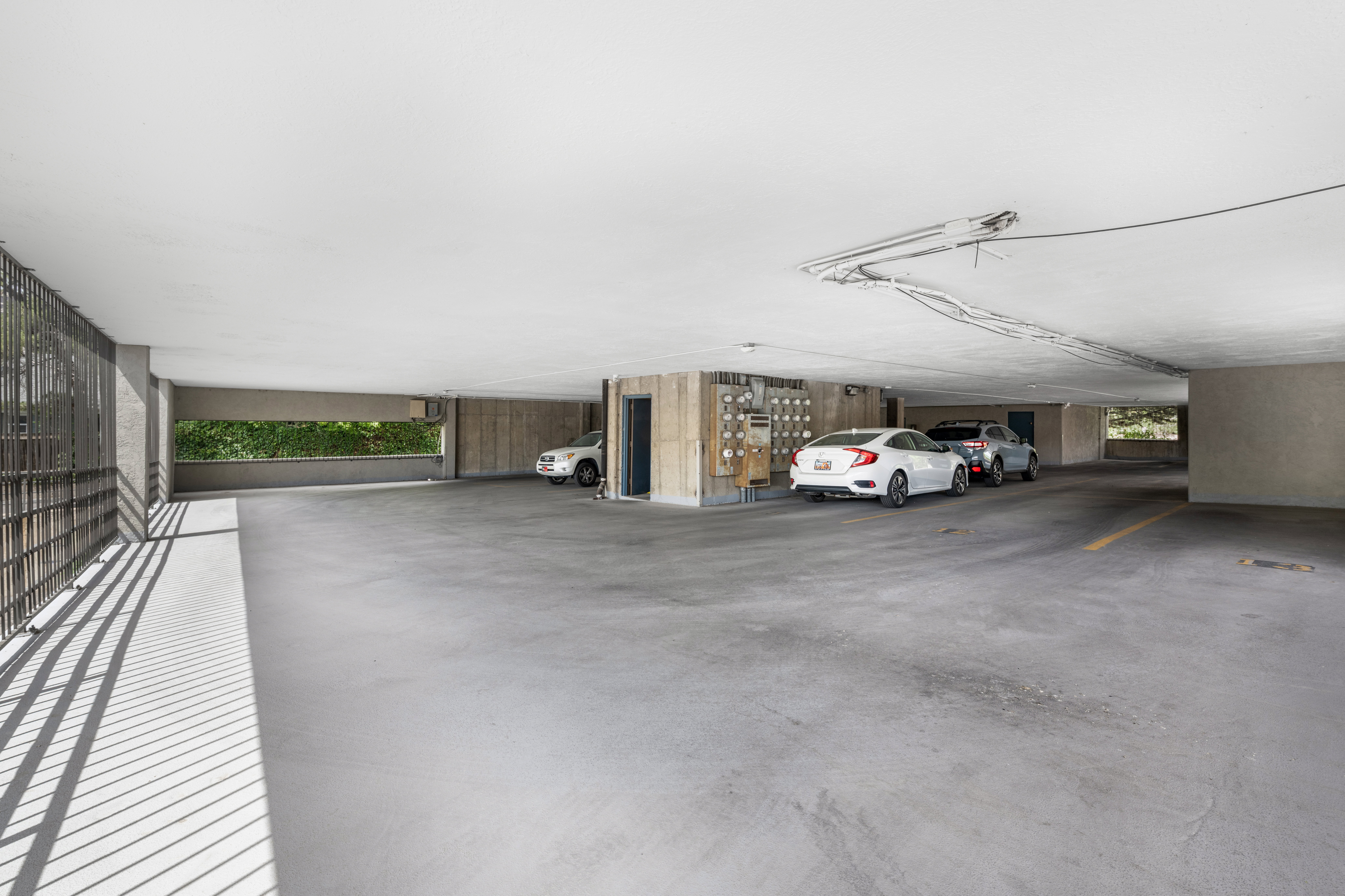 Concrete parking garage with multiple vehicle spaces, exposed ceiling conduits, and a decorative slatted metal partition