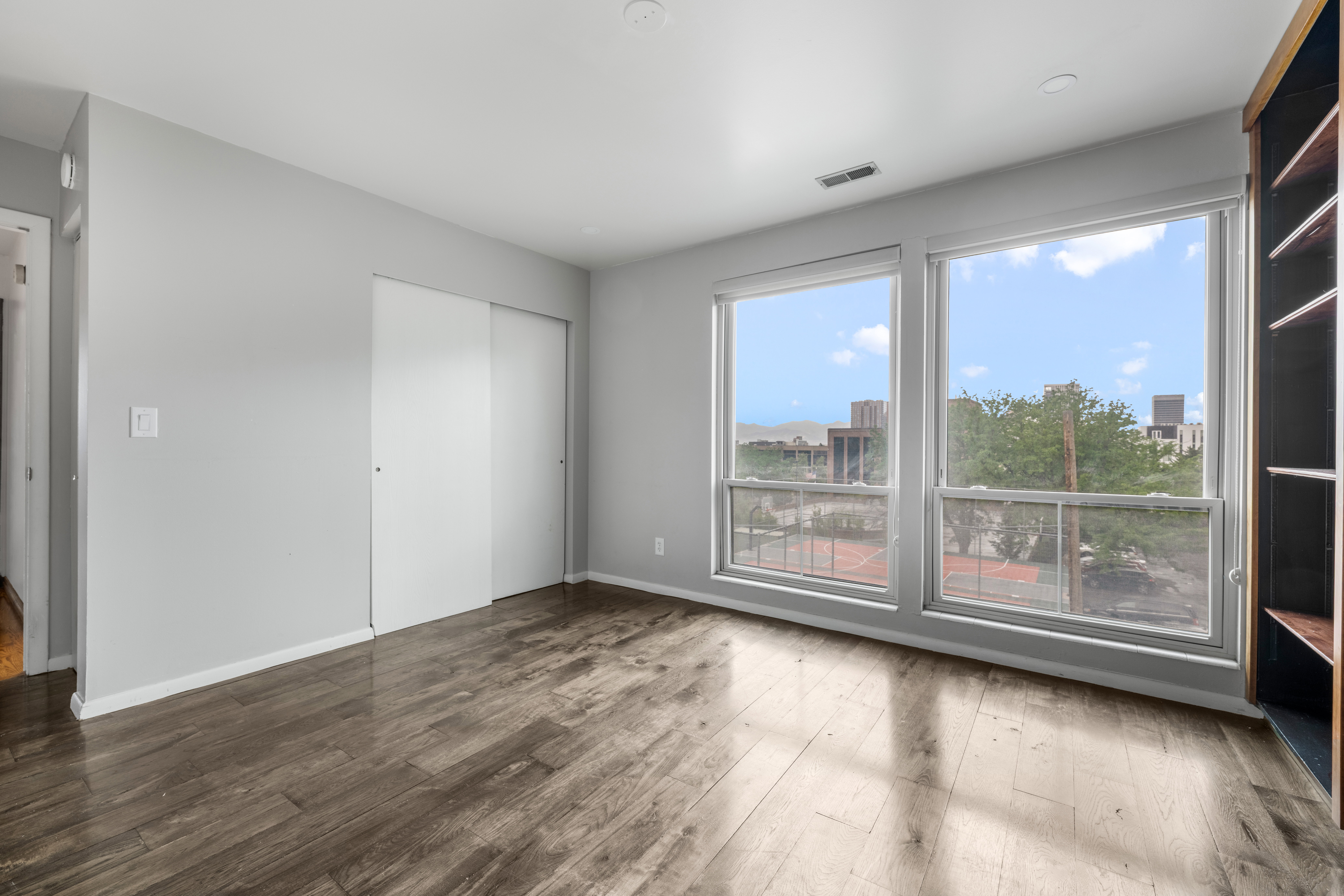 Spacious room featuring wood-finish flooring, light grey wall paint, two large windows, a built-in bookshelf, and sliding closet doors