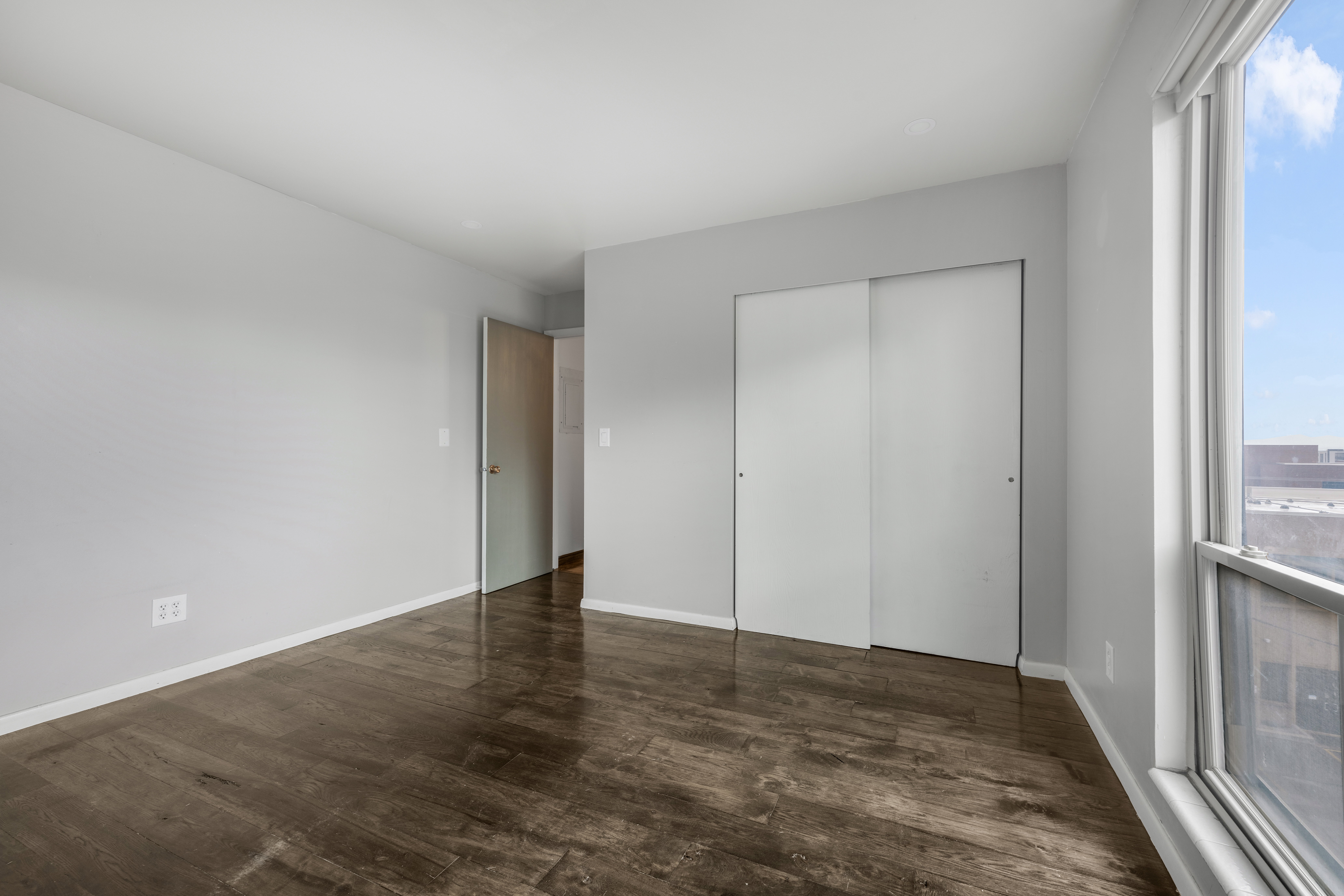 Spacious room featuring wood-finish flooring, light grey wall paint, white baseboards, and a large window providing natural light