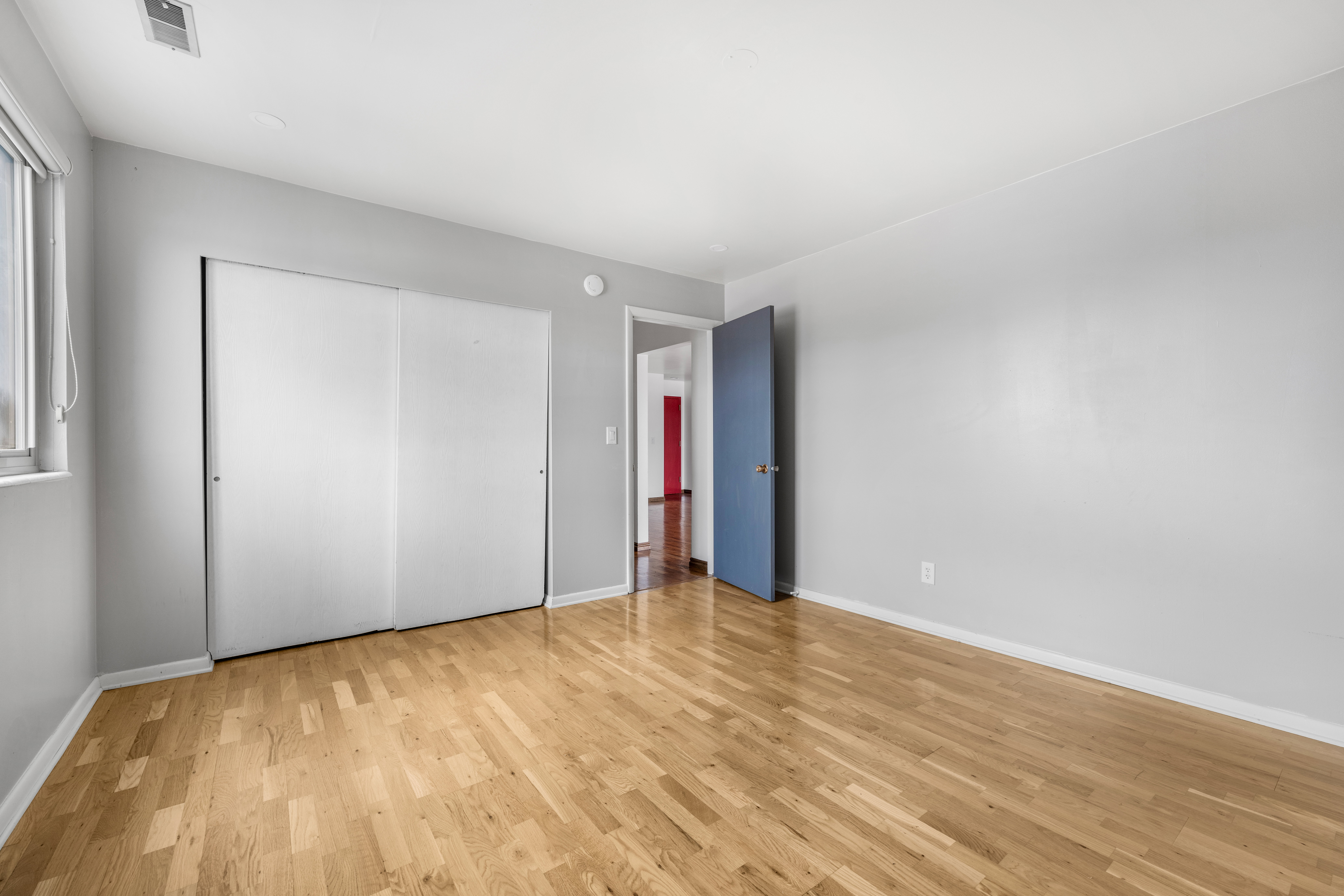 Hardwood flooring throughout featuring light gray walls, a window with blinds, and white bi-fold closet doors