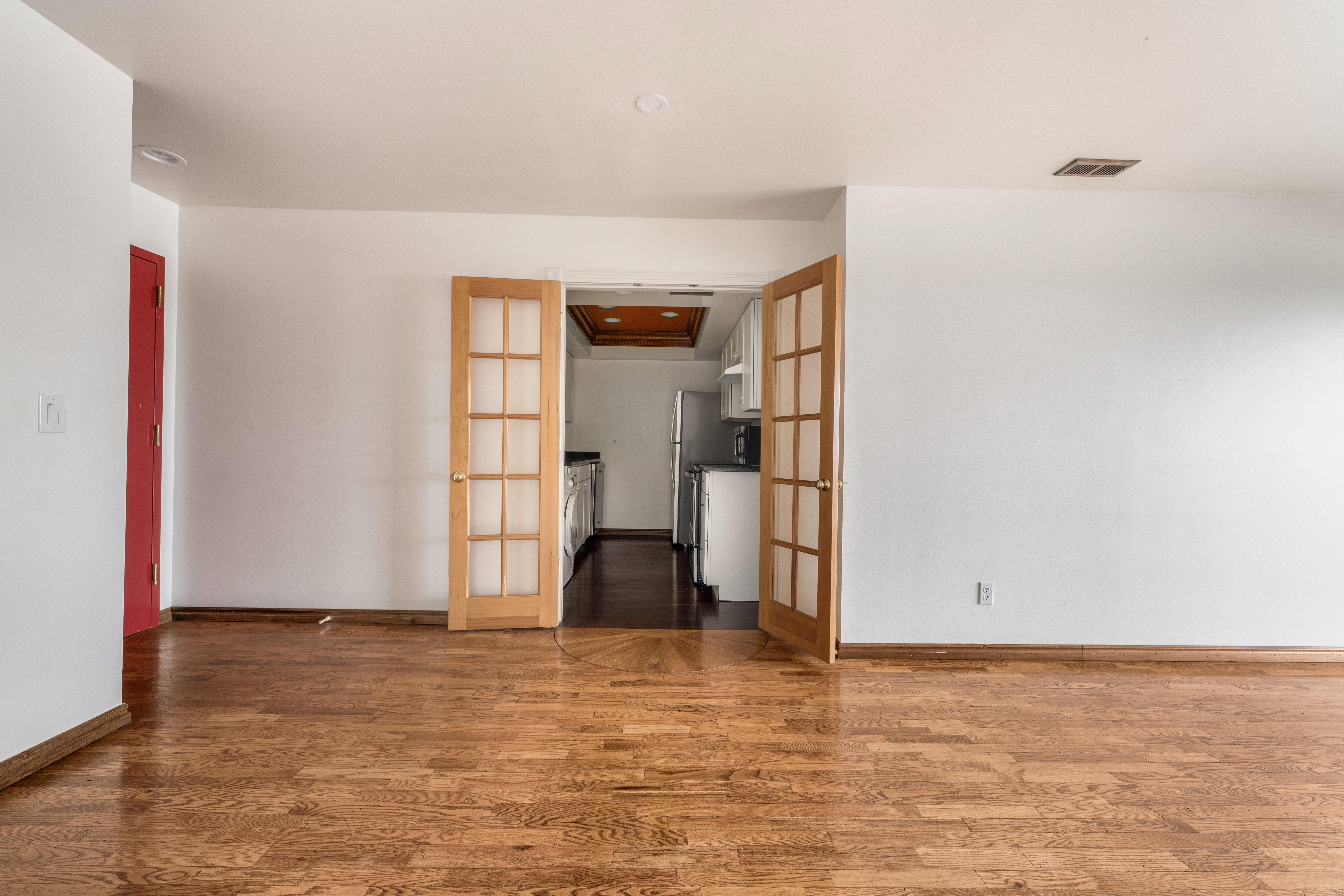 Hardwood flooring throughout a spacious room with white walls and a red entry door