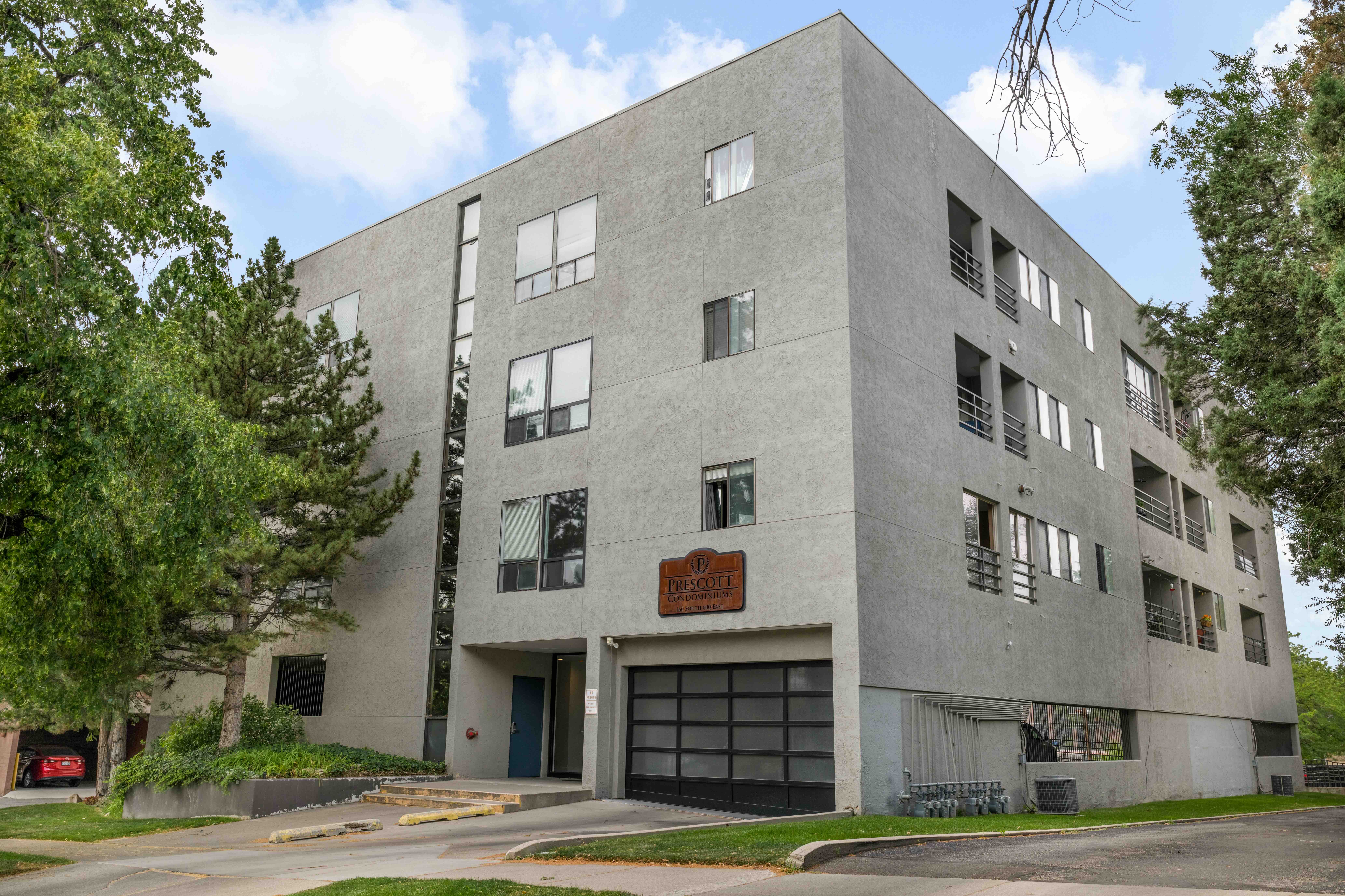 Contemporary multi-story building featuring a textured facade, floor-to-ceiling windows, private balconies with metal railings, and an integrated garage with a modern glass-paneled door