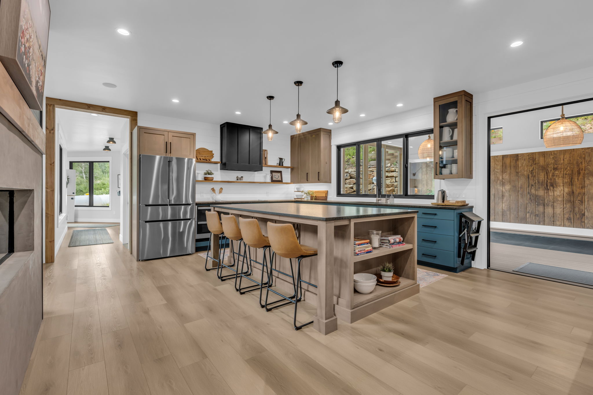Spacious kitchen featuring a large central island with integrated shelving, light wood-finish flooring, a stainless steel refrigerator, dark cabinetry, and multiple recessed light fixtures