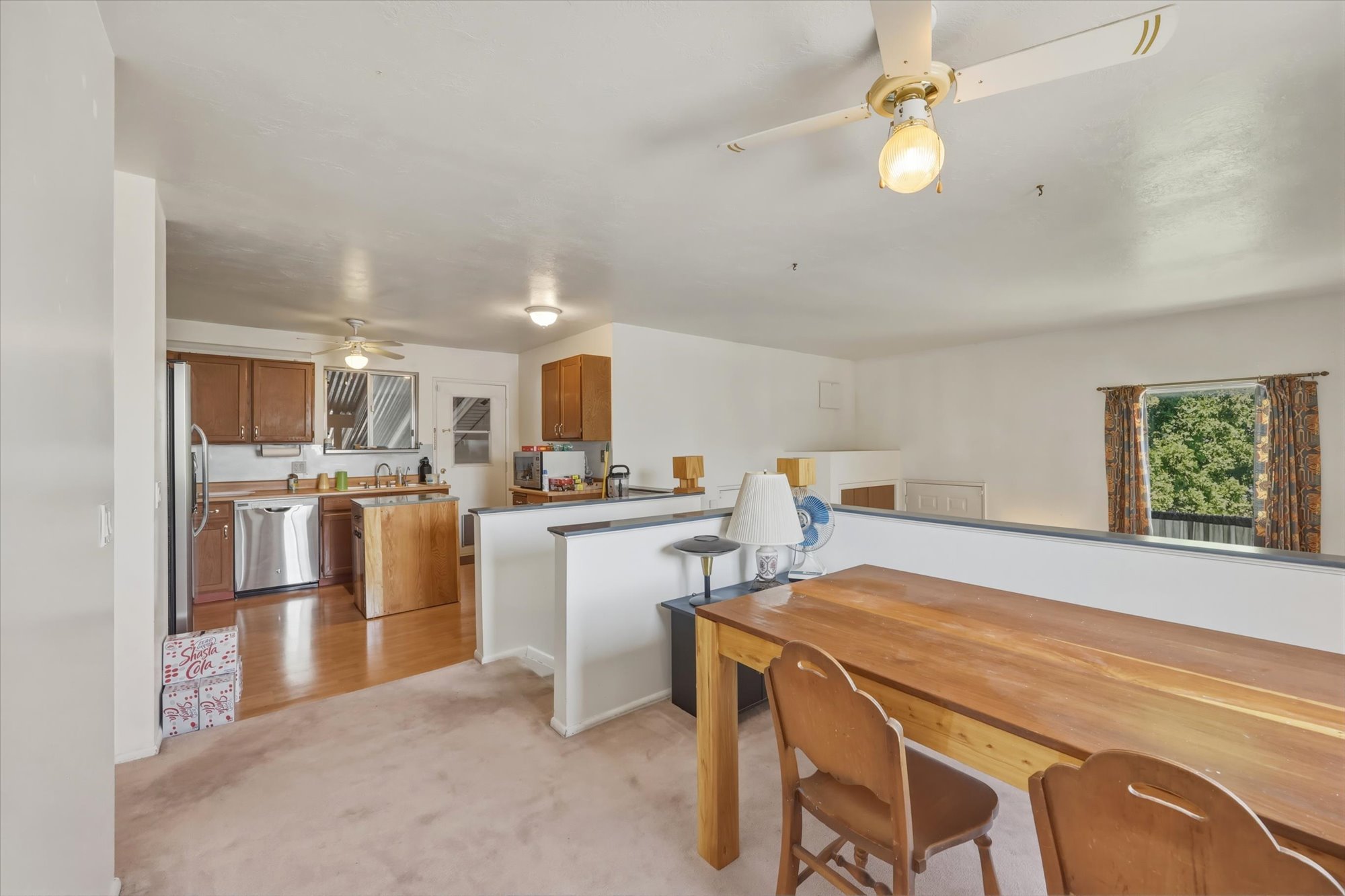 Bright interior with a ceiling fan, showcasing a kitchen with wood-finish cabinetry, stainless steel refrigerator, and a window with tree scenery