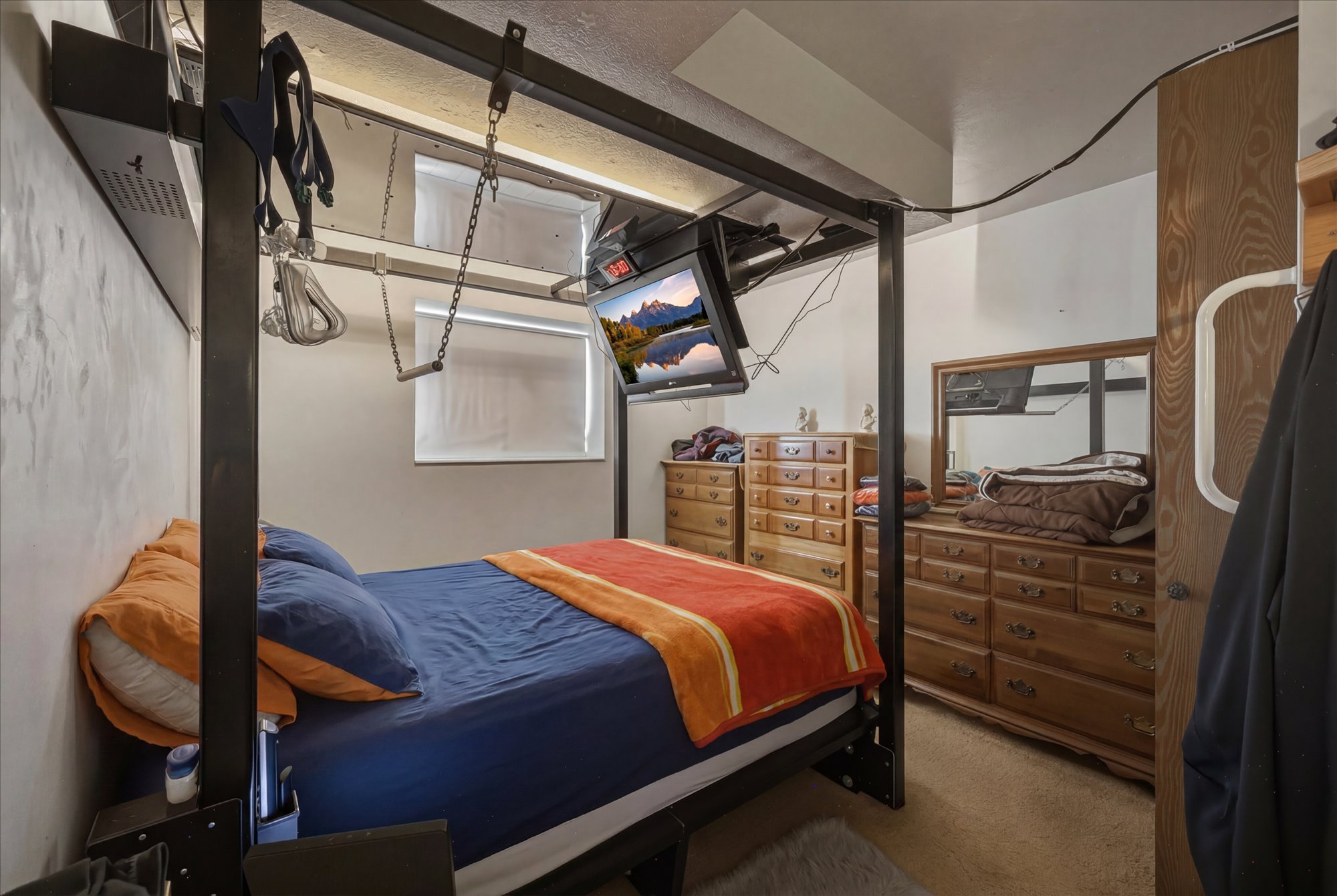 Carpeted room with a ceiling-mounted metal bed frame, featuring an integrated television and a window with a roller shade