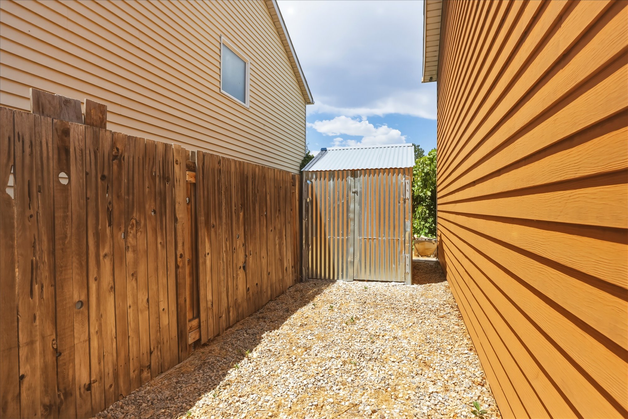 Gravel pathway featuring a corrugated metal shed with a matching roof, bordered by a wooden privacy fence and horizontal siding