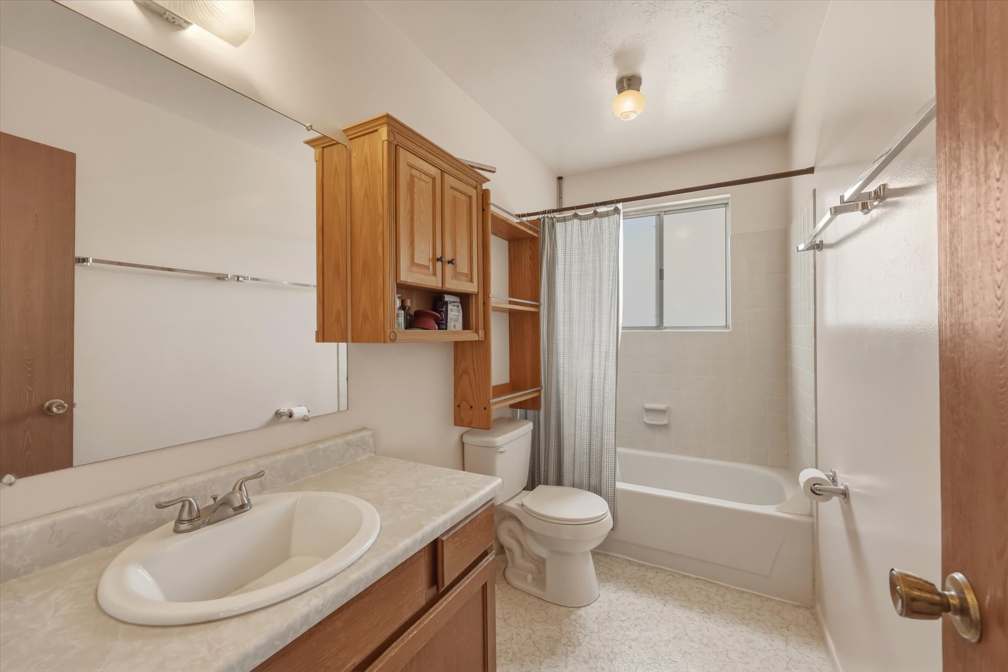 Full bathroom featuring a white bathtub/shower combination with tile surround, a single vanity with a laminate countertop, a large wall-mounted mirror, and wood-finish cabinetry providing storage