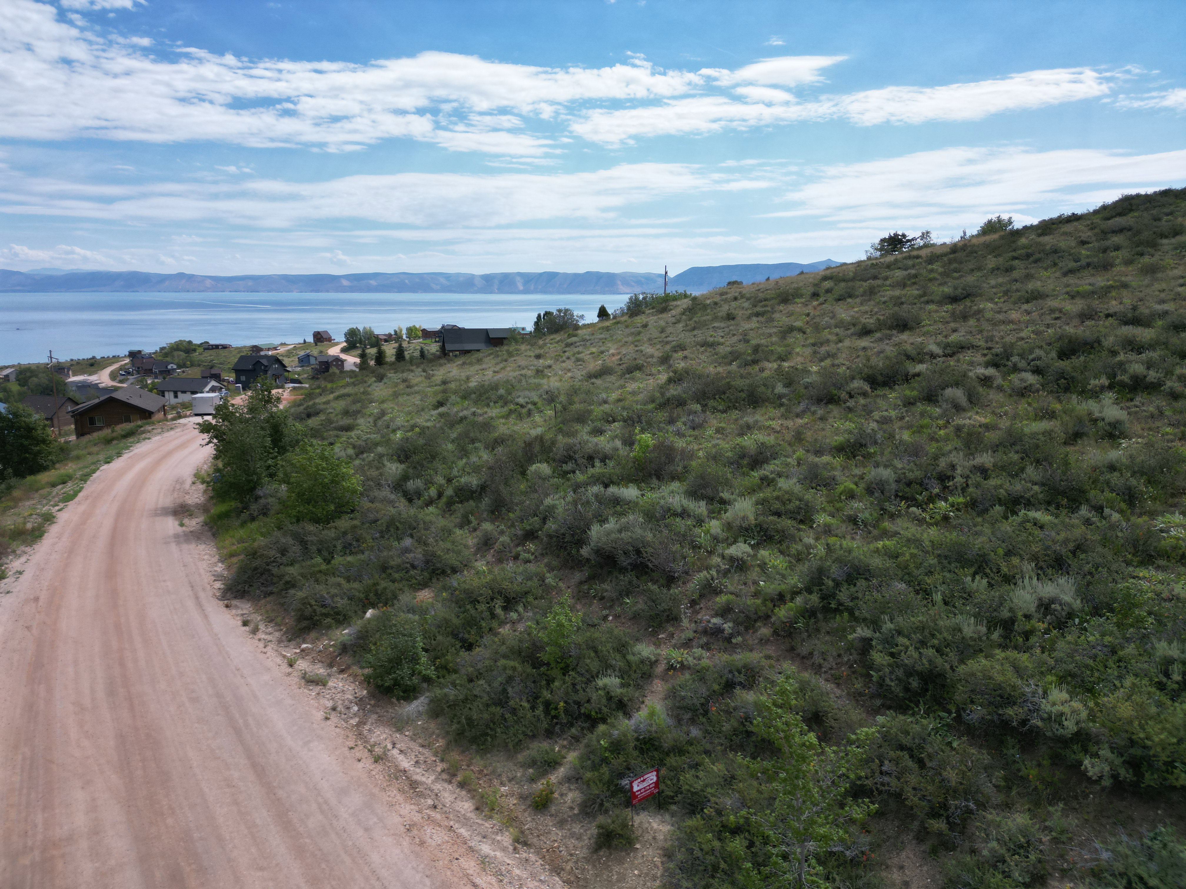 Expansive sloped terrain with natural shrubbery, featuring a visible dirt road, distant water body, and scattered residential structures
