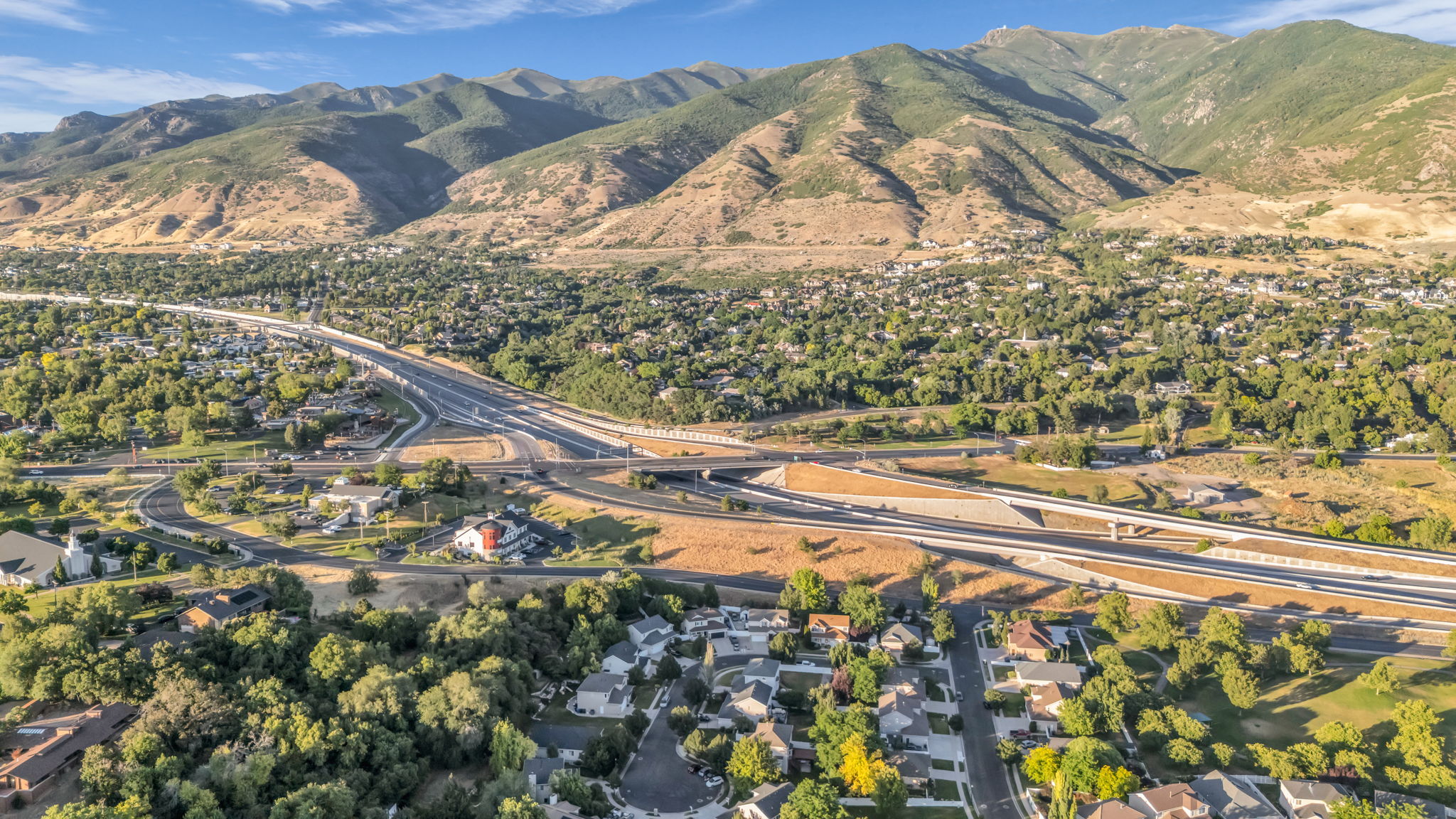 Expansive aerial perspective showcasing a mountainous backdrop, abundant green trees, dense residential areas, and prominent highway infrastructure