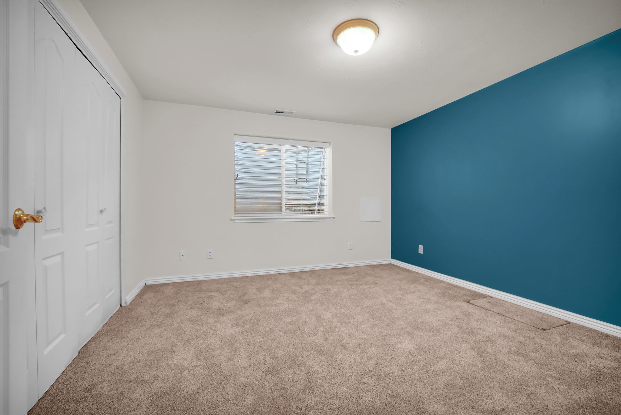 Carpeted interior space featuring a window with blinds, a prominent accent wall, and bi-fold closet doors with brass hardware