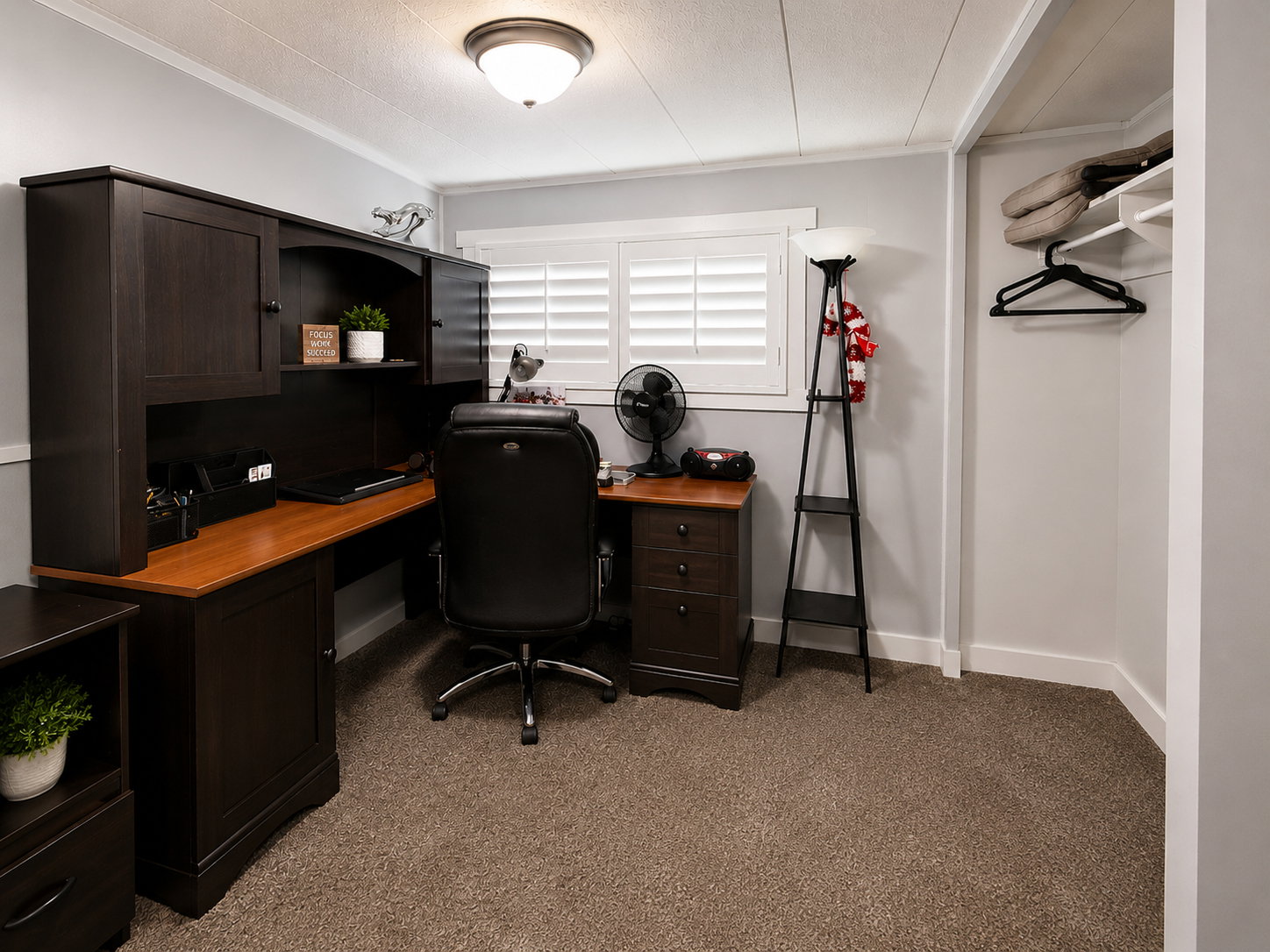 Dedicated office space featuring built-in dark wood-finish cabinetry, a window with plantation shutters, a ceiling-mounted light fixture, and plush carpeting