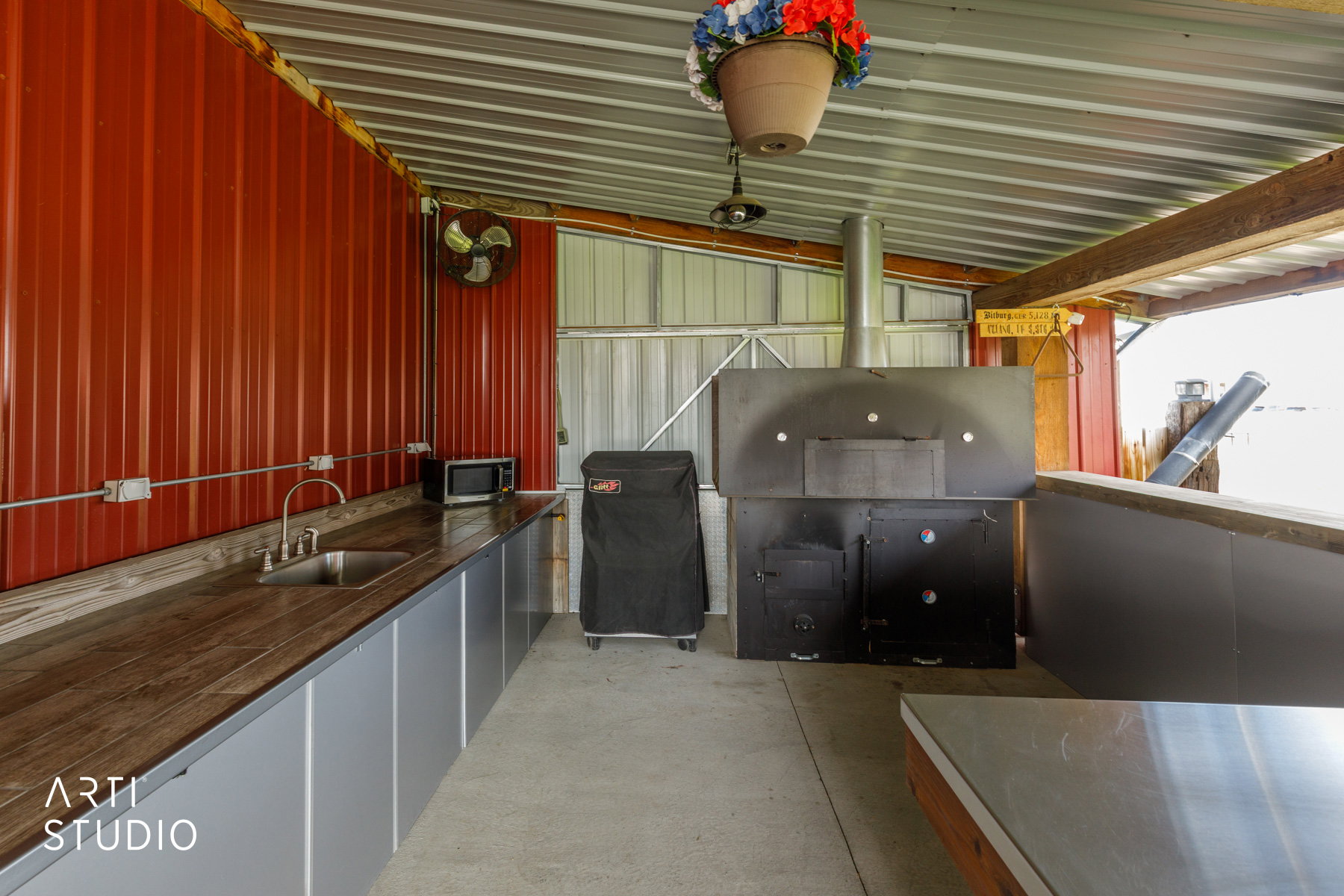 Covered outdoor kitchen featuring corrugated metal walls, wood-finish countertops, stainless steel sink, and a large built-in pizza oven with a metal flue