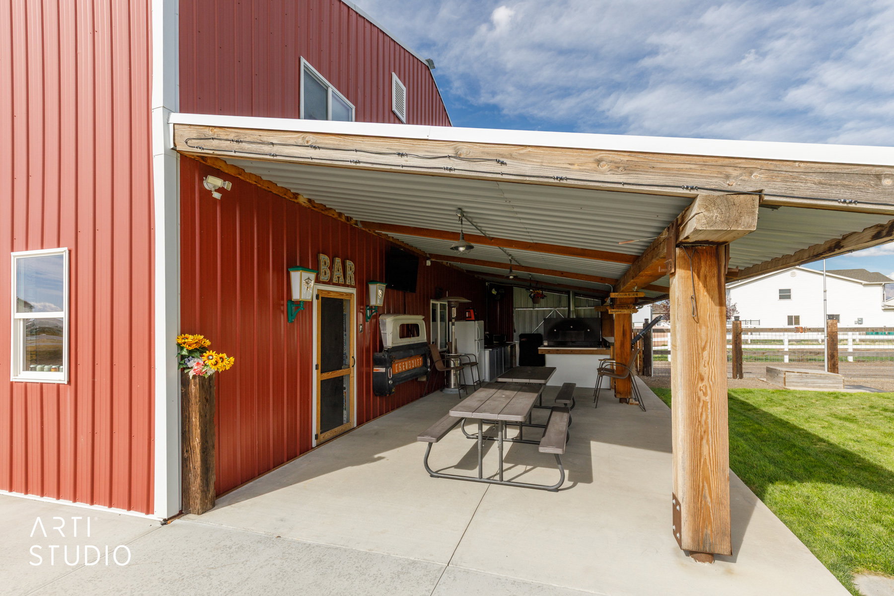 Expansive covered patio featuring a full outdoor kitchen, wood beam supports, concrete flooring, and corrugated metal roofing