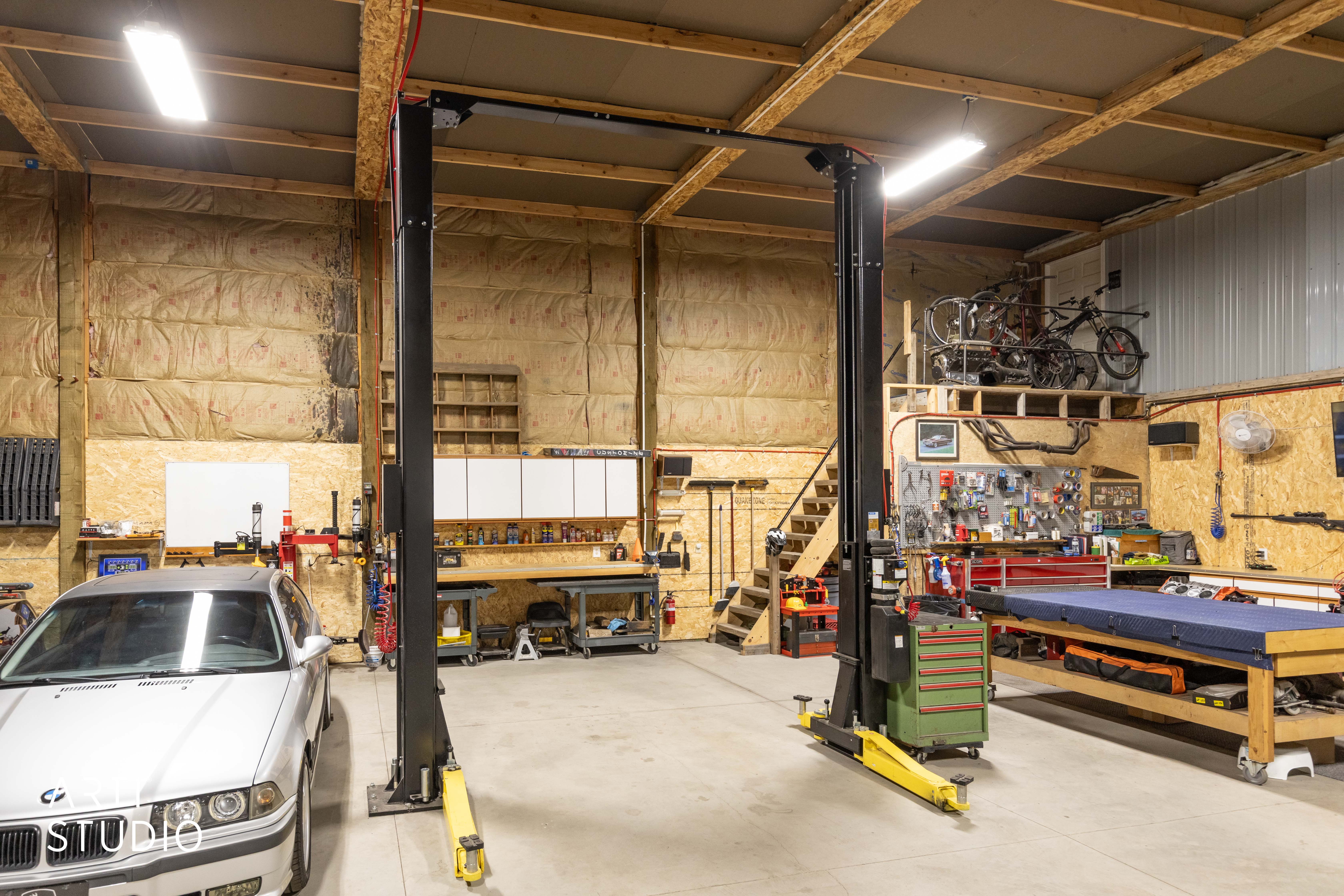 Spacious workshop featuring a hydraulic vehicle lift, exposed beam ceiling, concrete flooring, upper-level storage, and built-in cabinetry