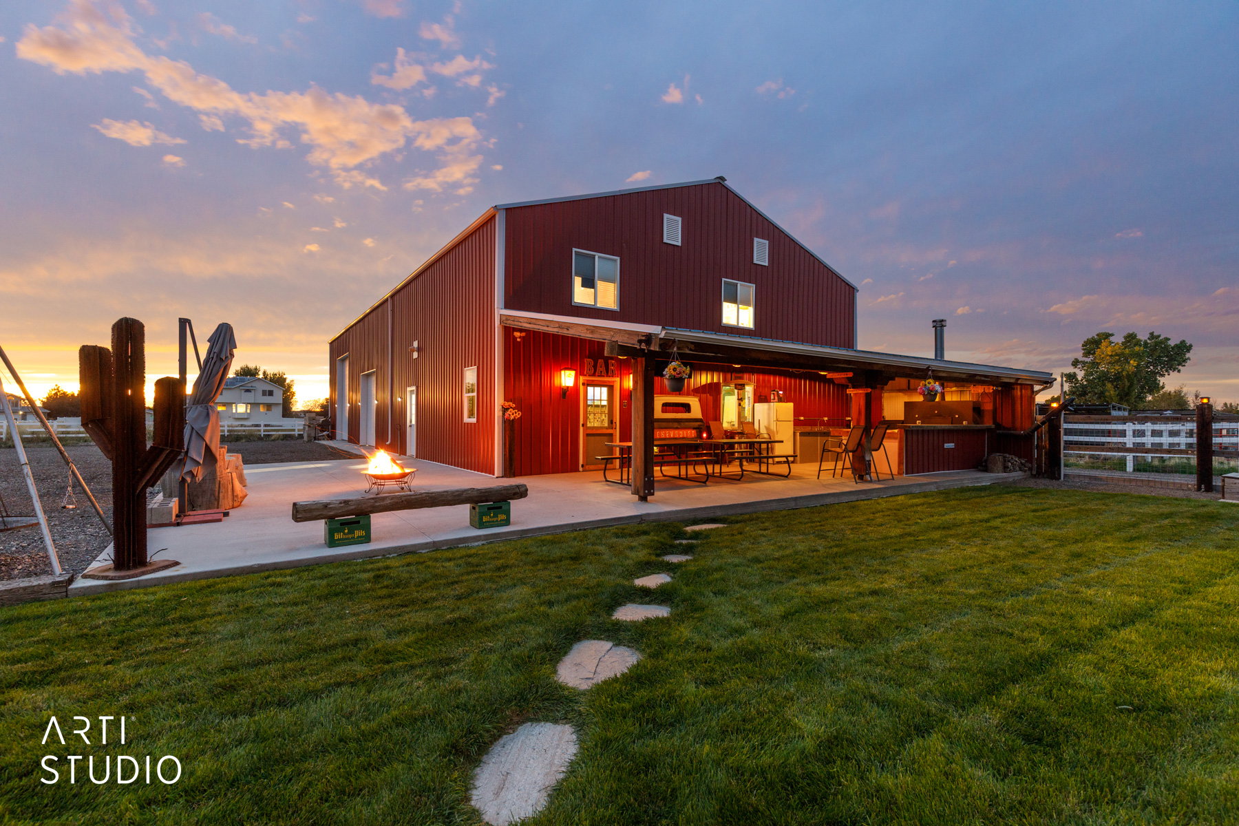 Red metal barn-style structure featuring a covered patio with an outdoor kitchen, a stone pathway, a lush green lawn, and a fire pit area
