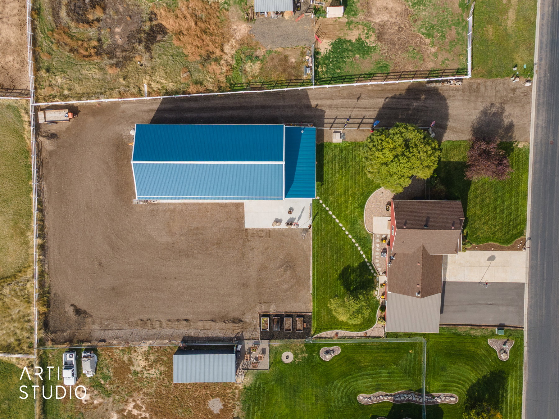 Aerial property overview featuring a large blue-roofed outbuilding, a red-sided residence with a brown shingle roof, and a substantial uncultivated land area