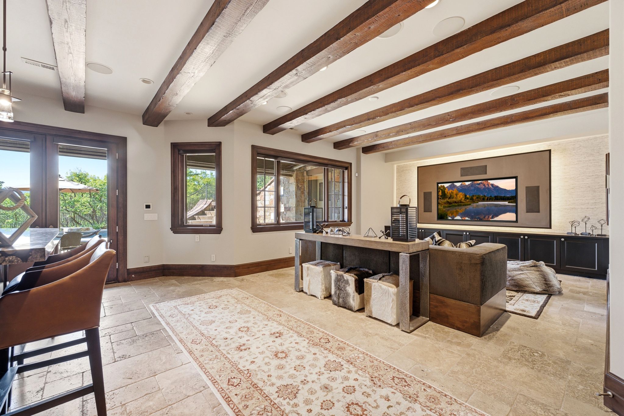 Expansive entertainment room featuring exposed wood ceiling beams, stone tile flooring, and a large built-in media wall with integrated speakers