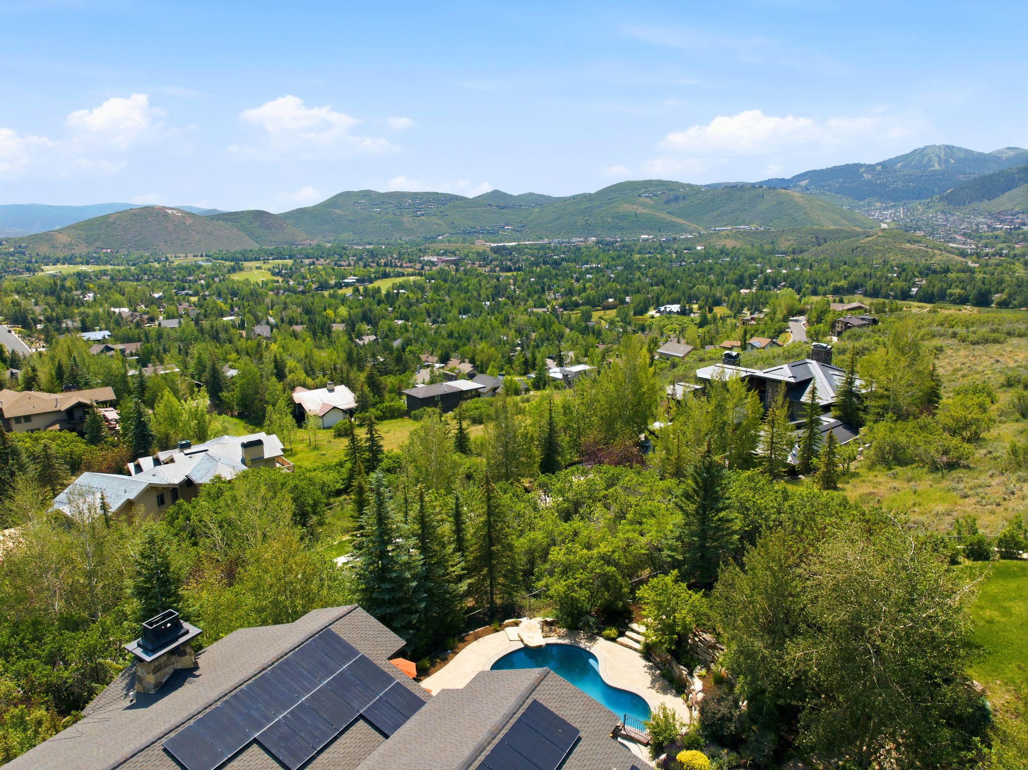 Aerial perspective showcasing a shingle roof with integrated solar panels, a sculpted swimming pool, and extensive surrounding greenery