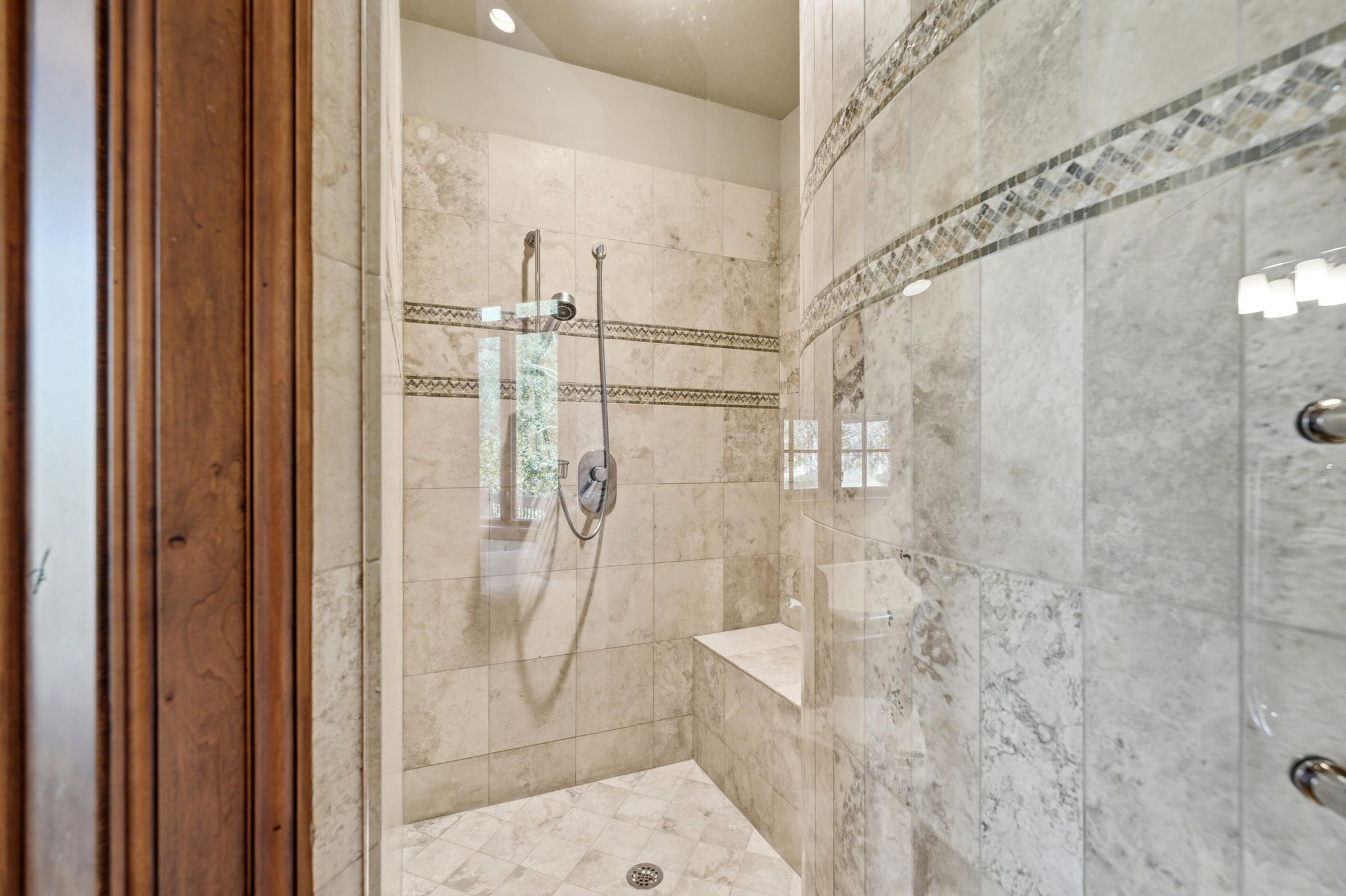 Walk-in shower featuring light-toned tile, a decorative mosaic tile band, a built-in bench, and a dual shower head system