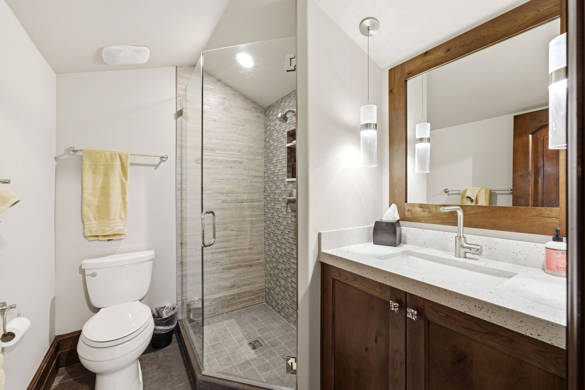 Contemporary bathroom featuring a glass-enclosed shower with tile surround and floor, a vanity with a light-toned countertop and integrated sink, a wood-framed mirror, and modern pendant lighting