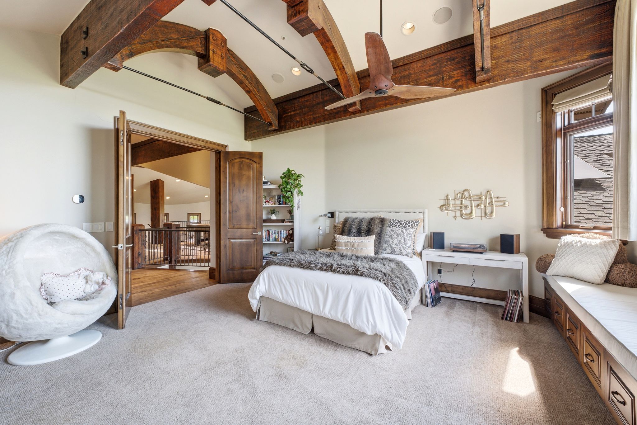 Vaulted ceiling with exposed wood beams and ceiling fan, wood-frame window with bench seating and integrated storage, double wood doors, built-in shelving, and carpeted flooring