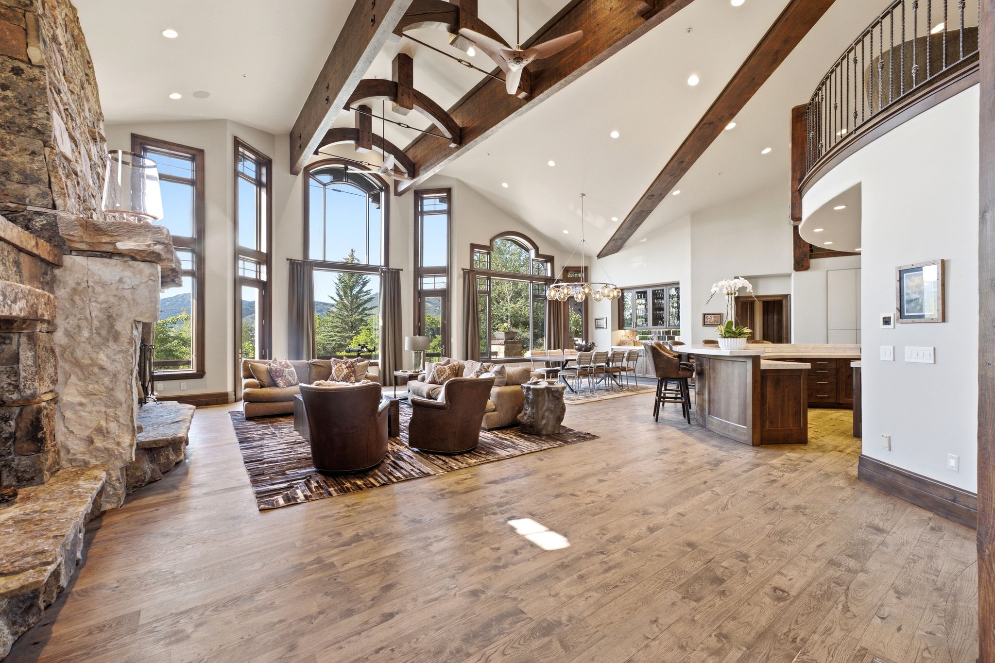 Great room featuring vaulted ceilings with exposed wood beams, a stone fireplace, wood-finish flooring, a curved upper balcony with wrought iron railing, and expansive windows