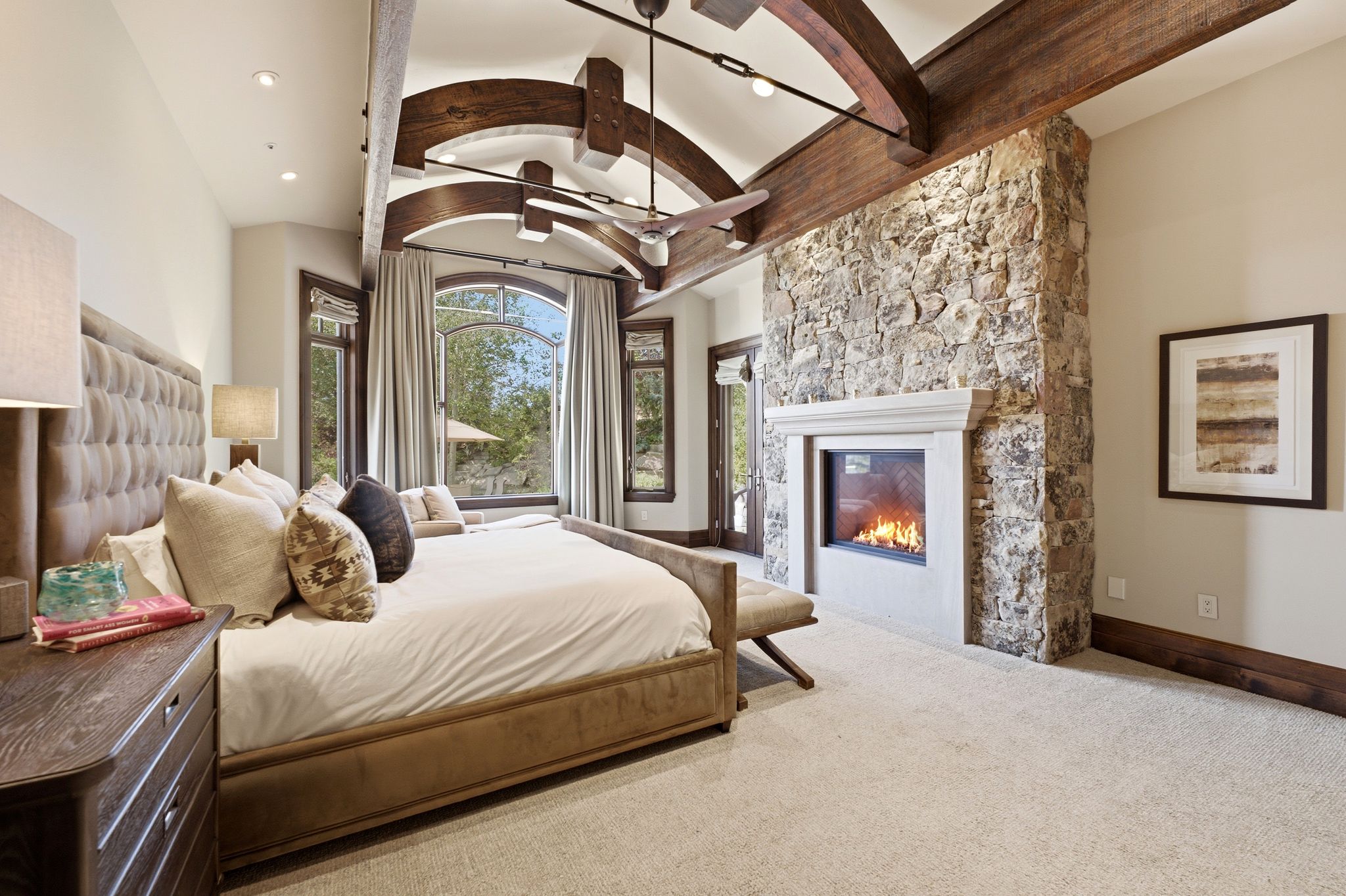 Carpeted interior space featuring a stone-clad fireplace with a built-in firebox, exposed wood ceiling beams, and an arched window with dark wood framing