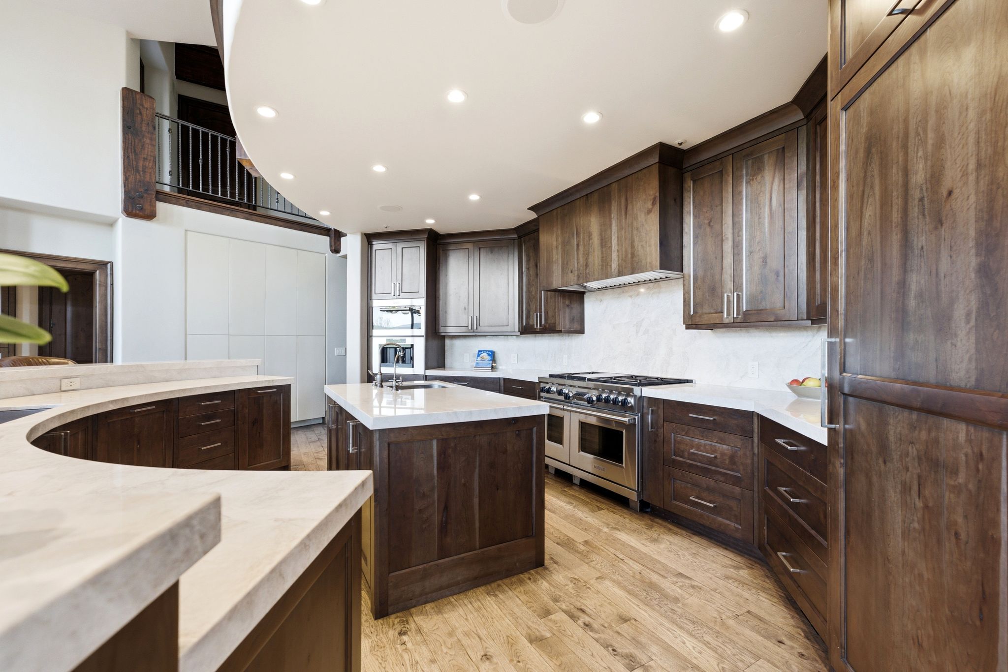 Gourmet kitchen featuring a large central island, curved breakfast bar, custom wood cabinetry, light-toned countertops, and a professional-grade stainless steel range with double ovens