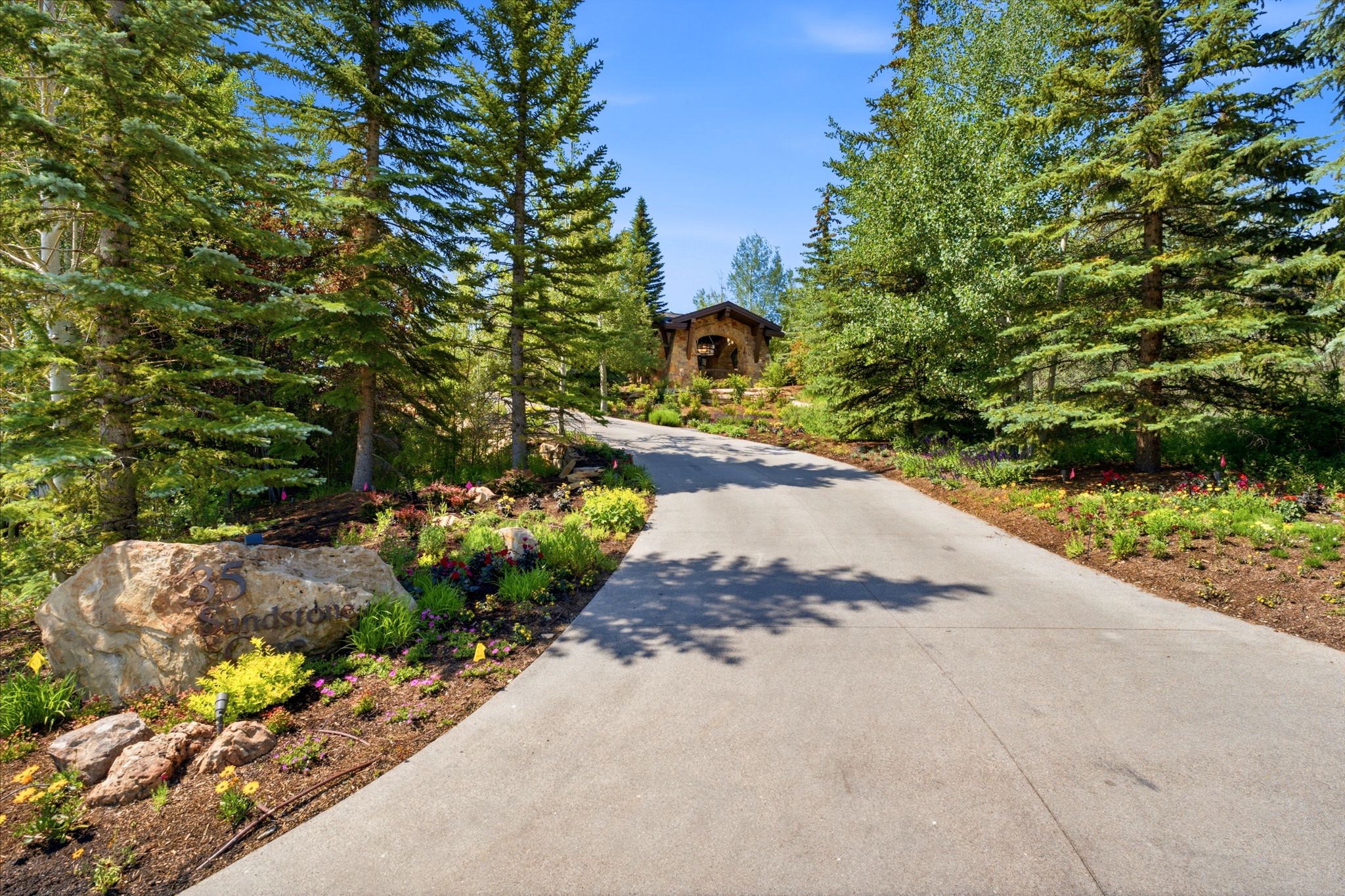 Paved driveway with lush evergreen and deciduous trees, bordered by landscaped natural rock and plantings