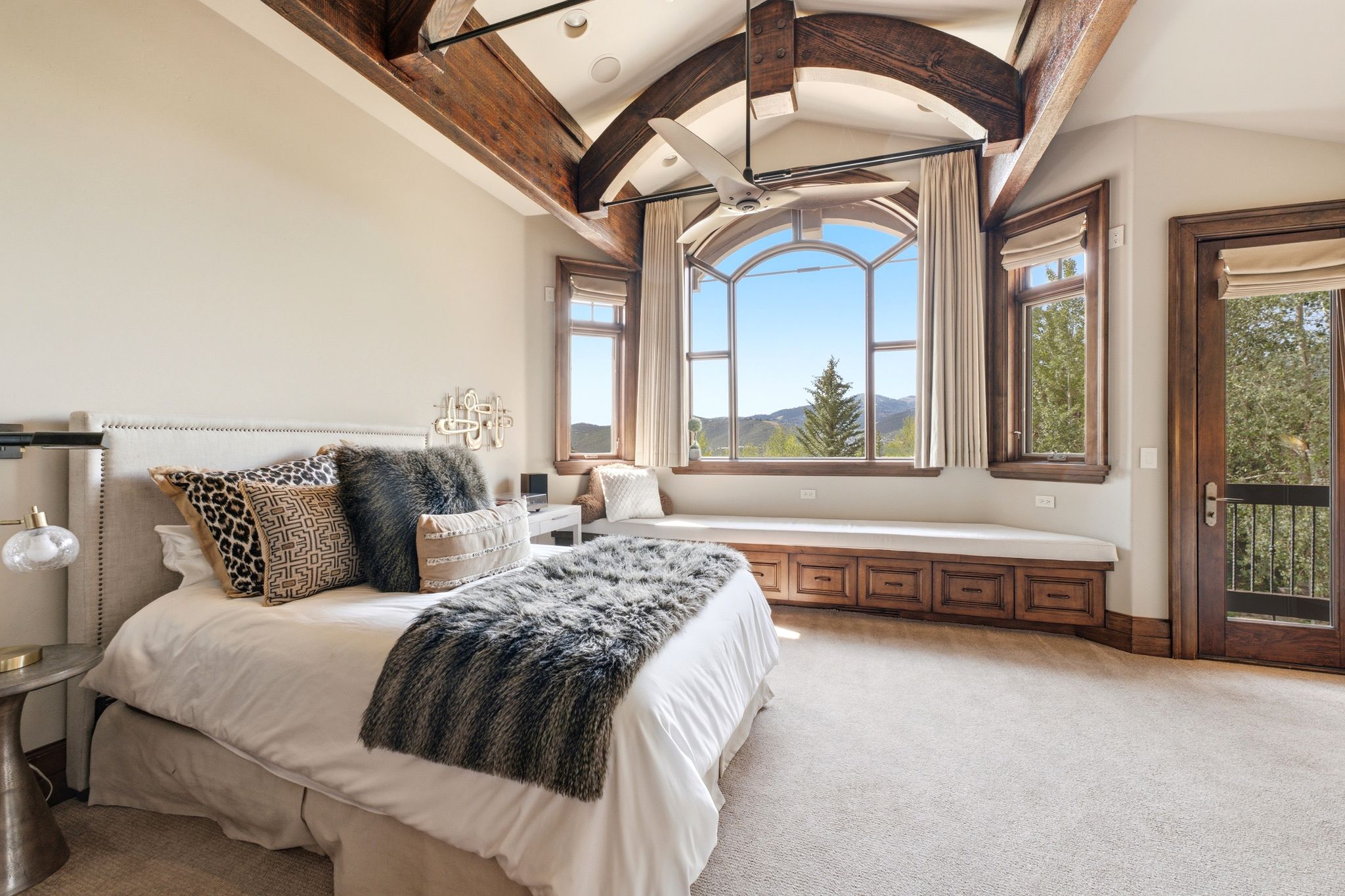 Vaulted ceiling with exposed wood beams, arched picture window, integrated window bench with storage, ceiling fan, and wood-paneled door with wrought iron balcony railing